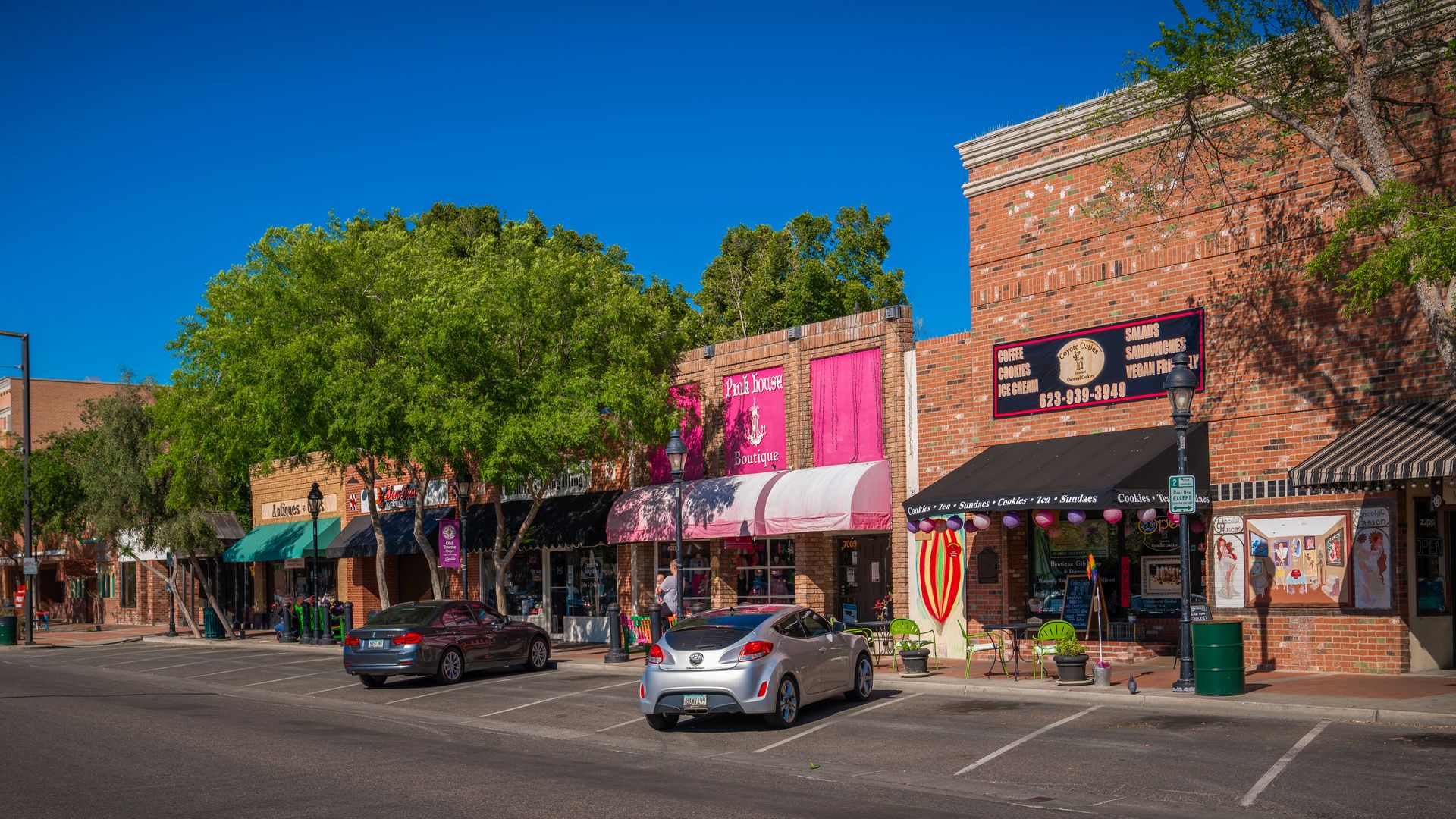 Bright street scene with brick storefronts, green trees along the sidewalk, and a row of shops under a vivid blue sky. Pink awnings and banners, outdoor seating, and parked cars line the curb.