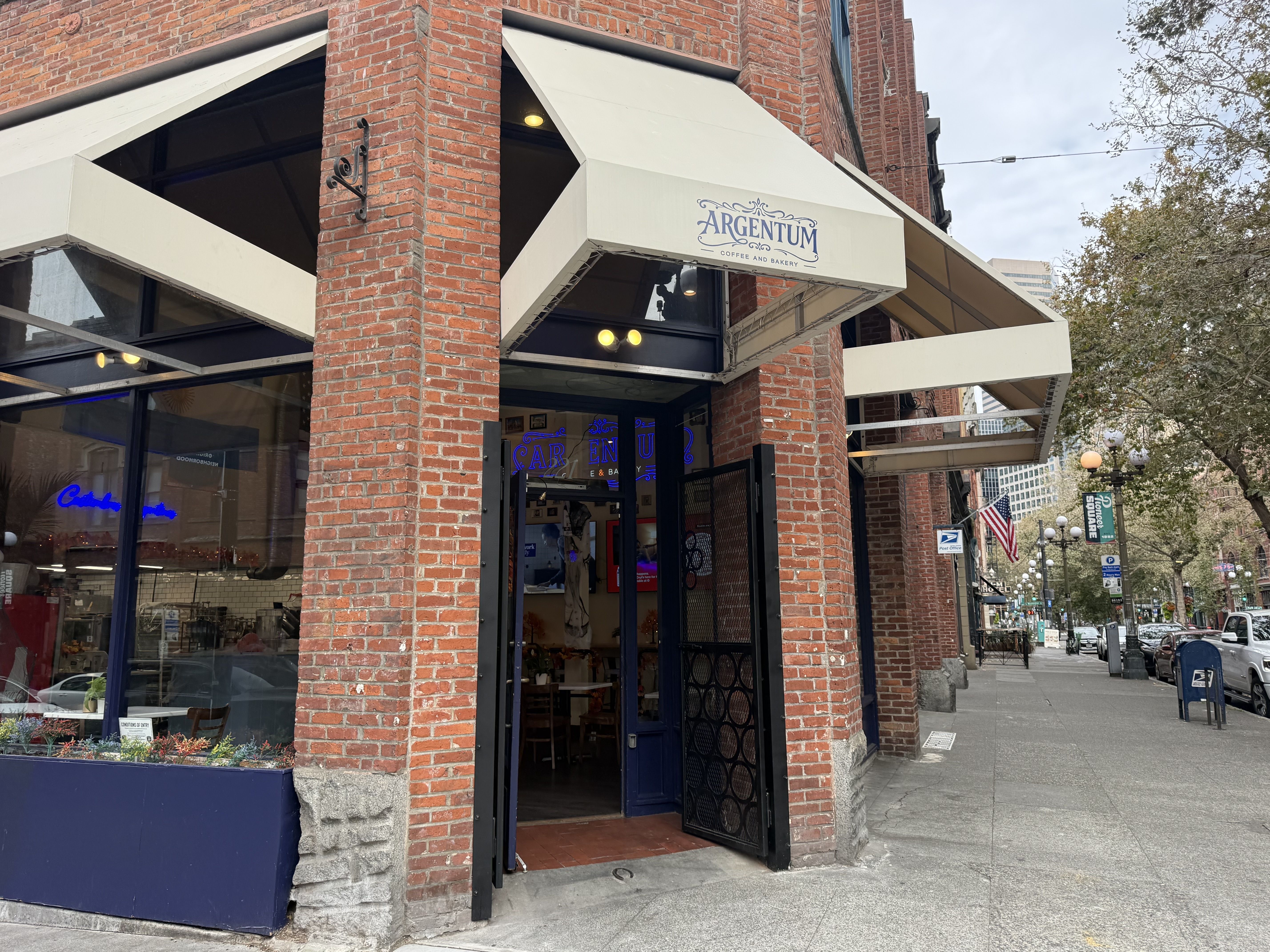 Brick corner cafe with white awnings and a blue doorway; sign reads Argentum Coffee and Bakery. Open entrance, glass storefronts, street scene with trees and flags in the background.