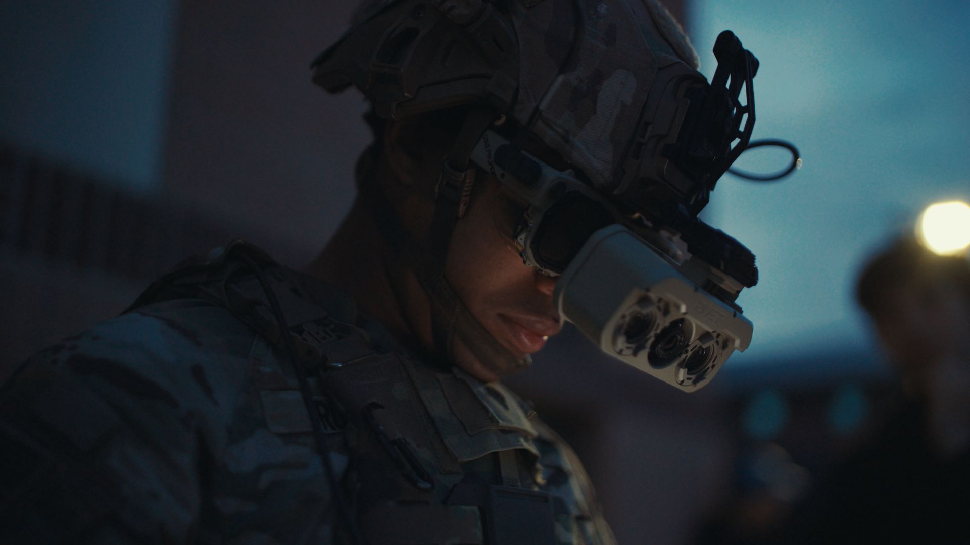 Side profile of a camouflaged soldier wearing a helmet with a futuristic headset in a dim blue-lit setting.
