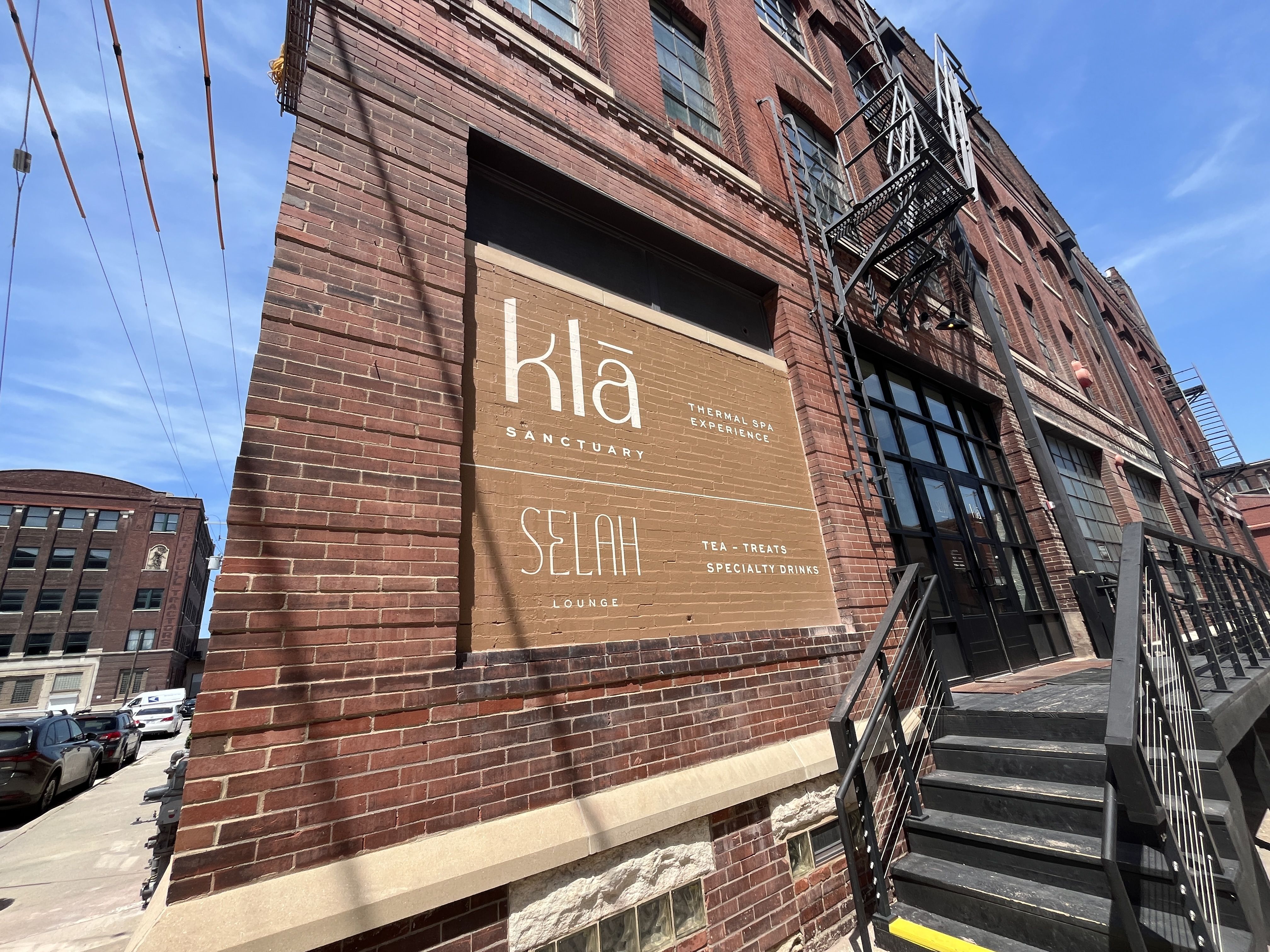 Brick building with a brown sign reading "klā Sanctuary Thermal Spa Experience" and "Selah Lounge Tea, Treats, Specialty Drinks" by black metal stairs under blue sky.