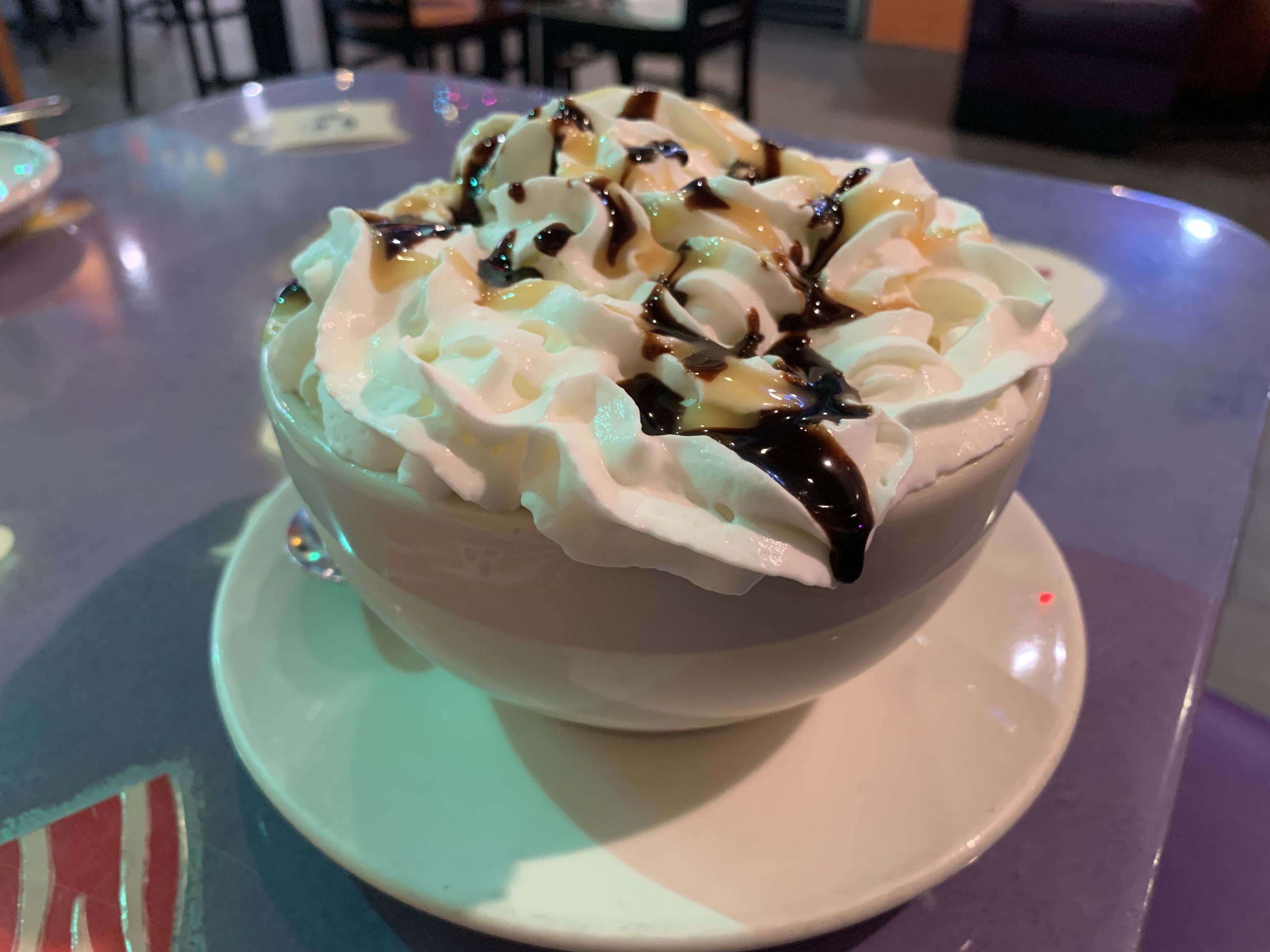 White bowl on a saucer filled with whipped cream topped with chocolate and caramel syrup, placed on a purple table in a dimly lit setting.