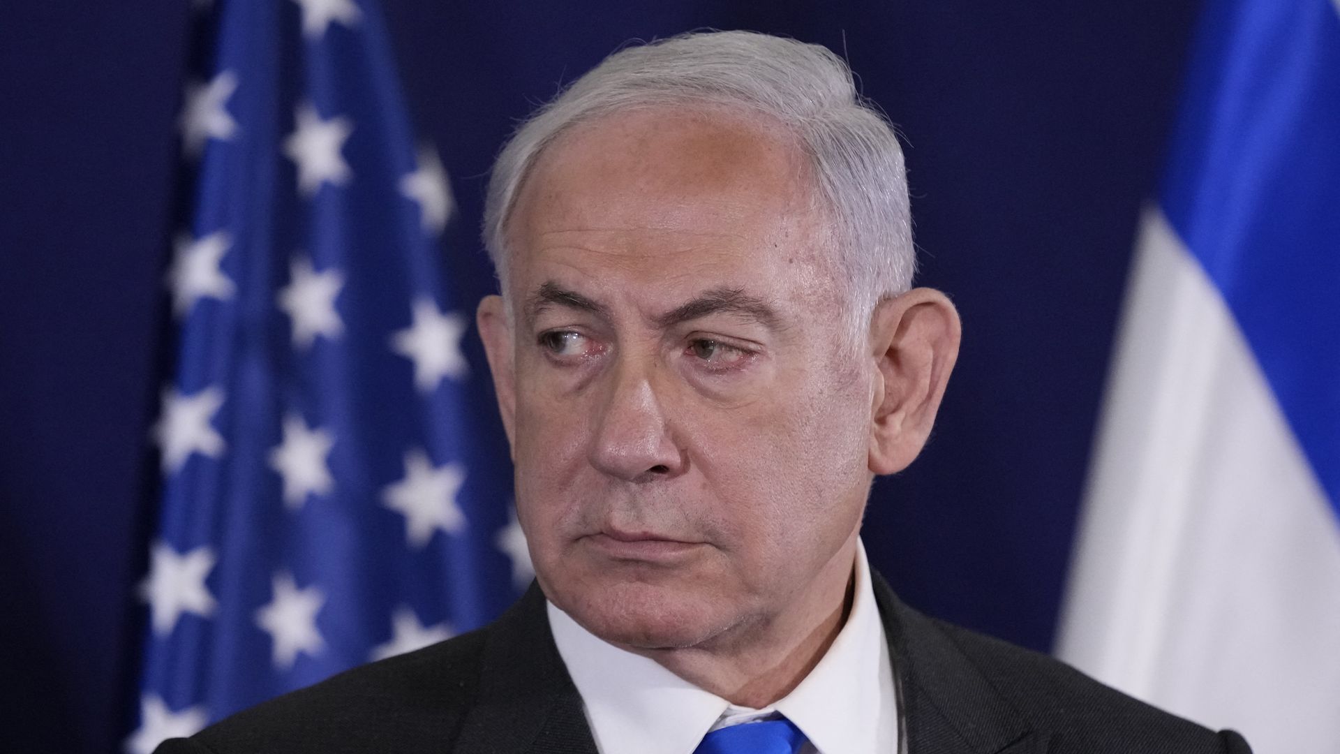 Israeli Prime Minister Benjamin Netanyahu, wearing a dark gray suit, white shirt and blue tie, standing in front of a U.S. and Israeli flag.