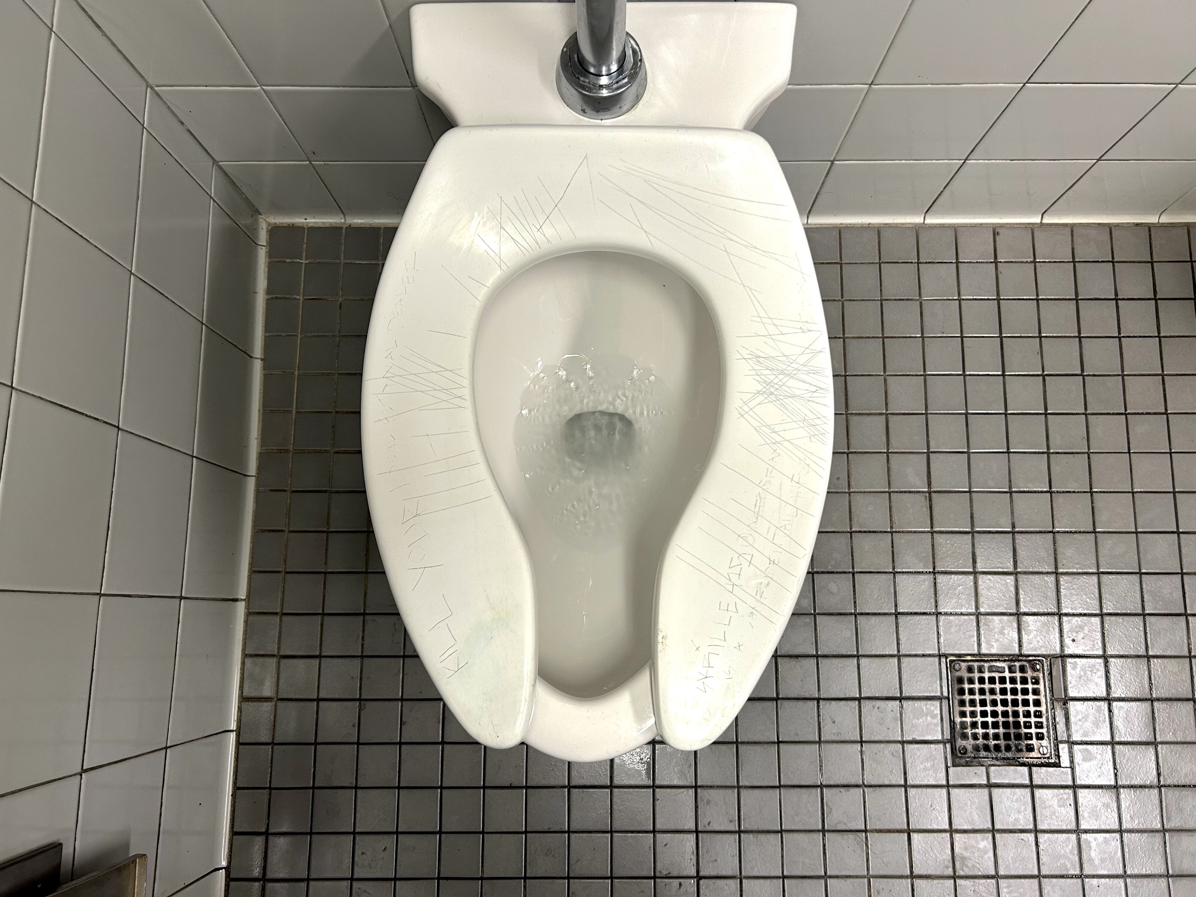 Top-down view of a white toilet in a gray tiled bathroom. The seat is scratched on both sides; a floor drain appears to the right and a chrome flush handle sits behind the bowl.