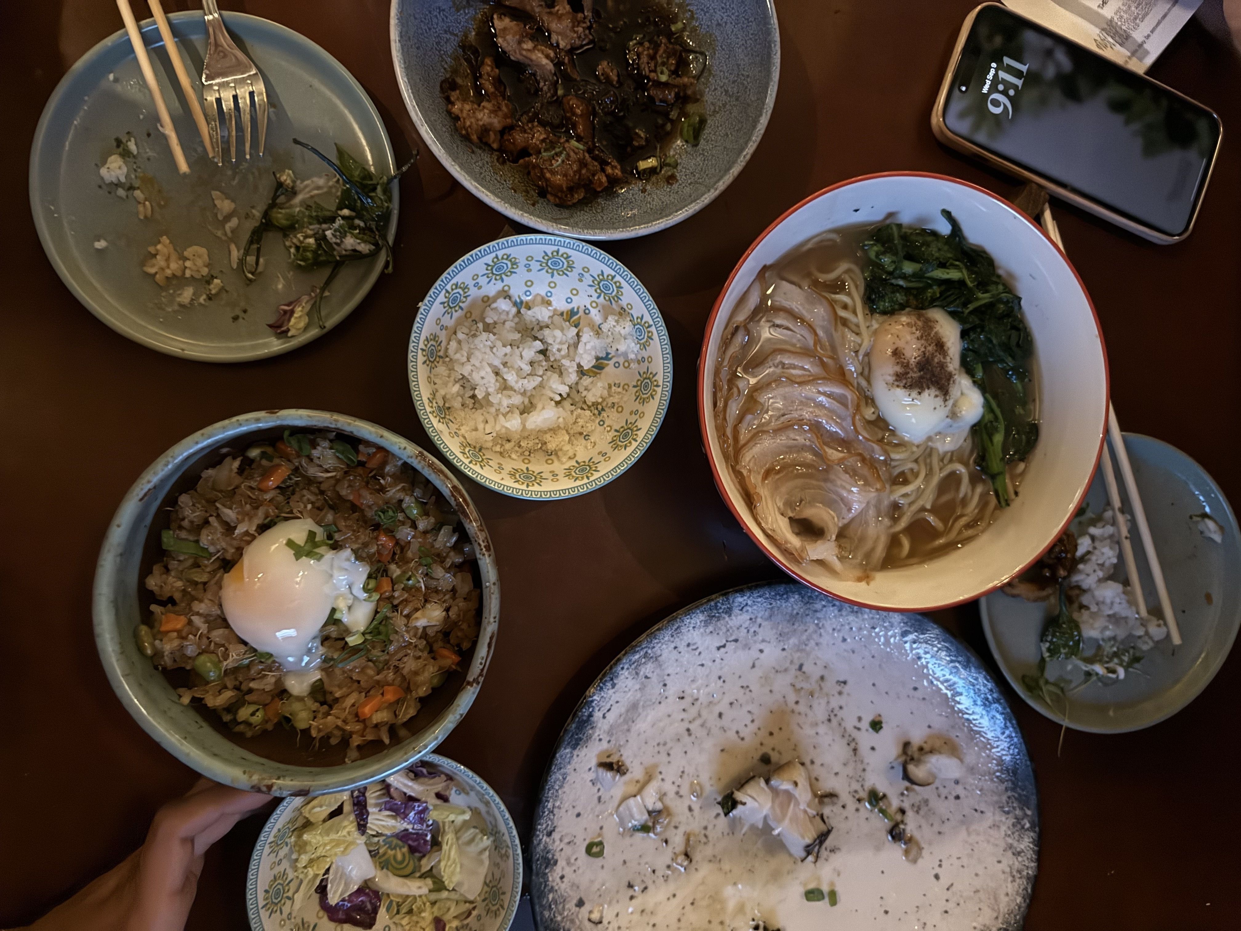 Overhead view of a table with several bowls of food: noodle soup with greens and a soft-boiled egg, fried rice with egg, a saucy meat dish, chopsticks, and a smartphone on the right.