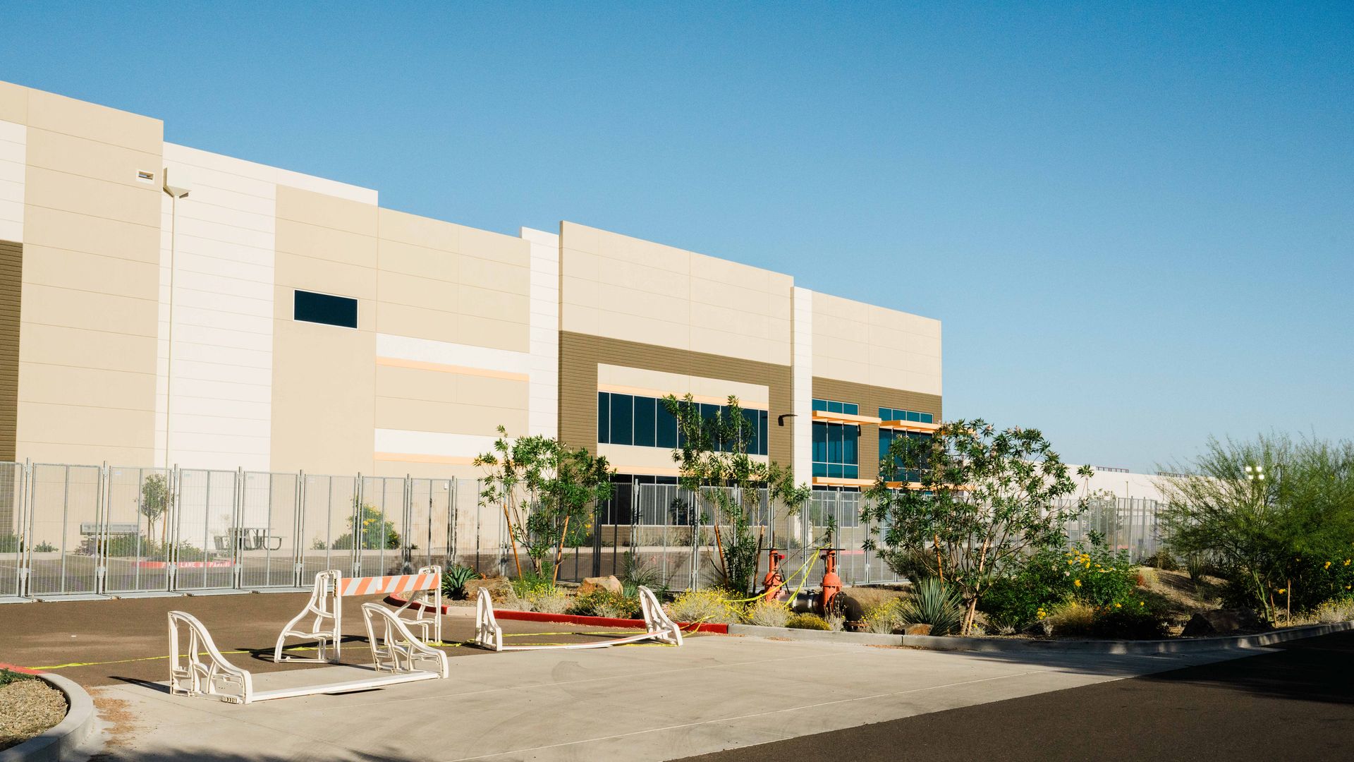The exterior of a beige and white warehouse surrounded by temporary chain-link fencing; several trees and construction-style barricades are outside the fence. 