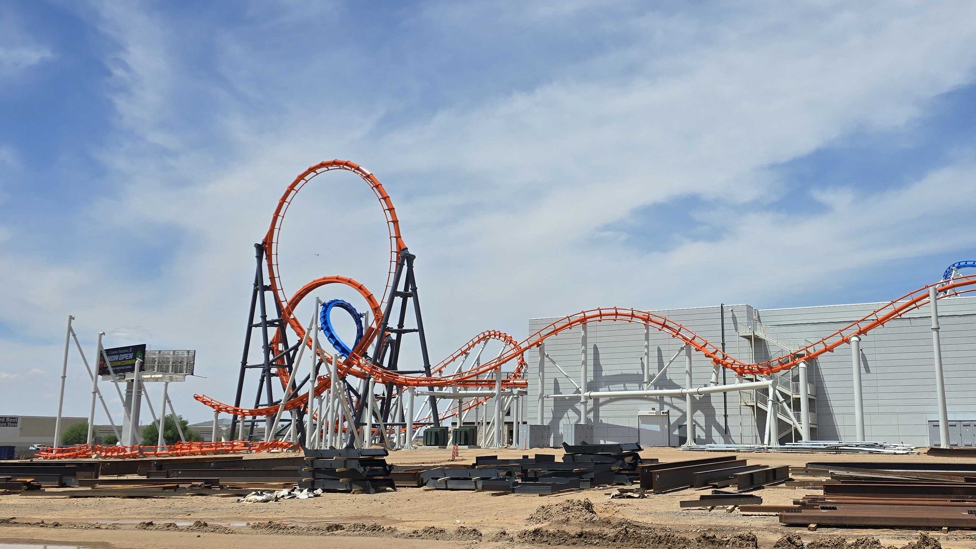 Construction site with orange roller coaster track looping over dark gray supports, blue track segments to the right, a white industrial building in the background, under a sky with wispy clouds.