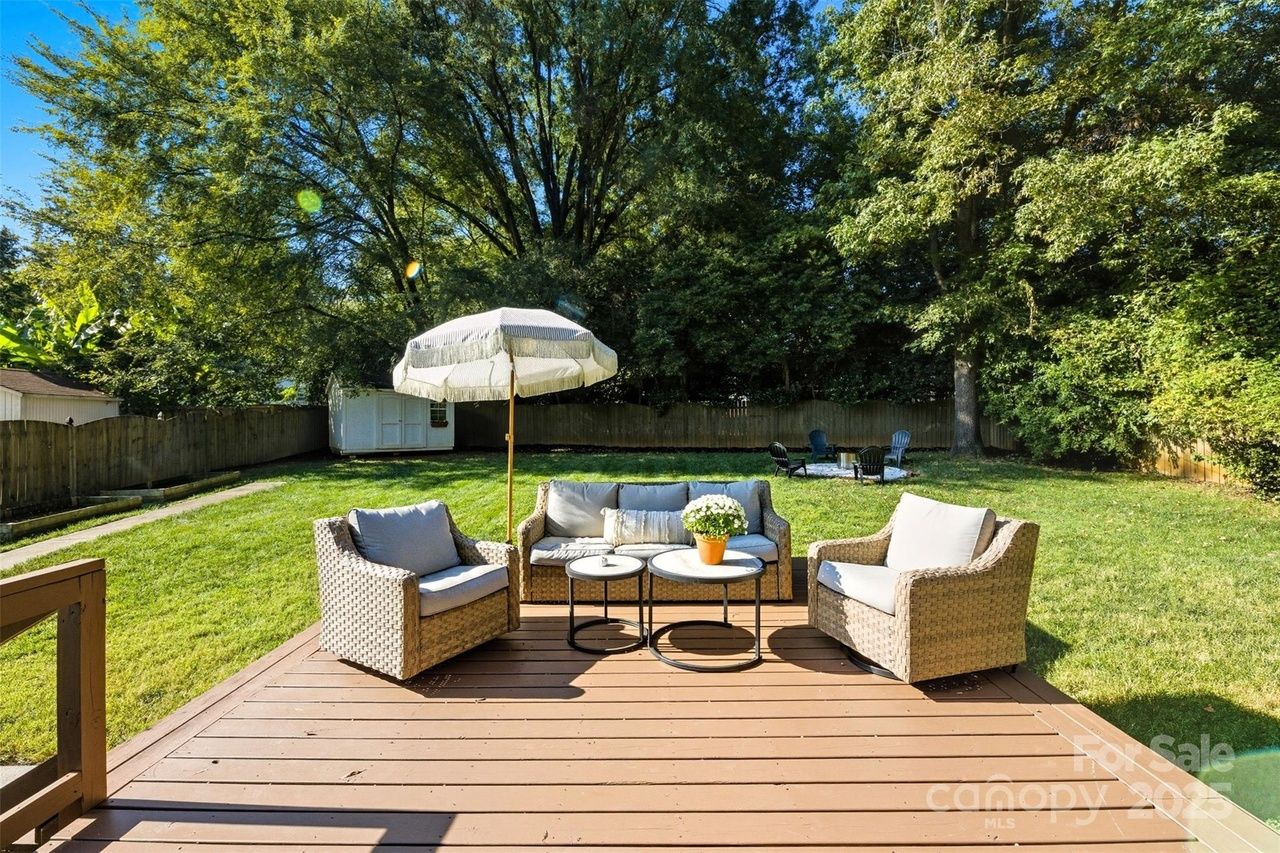 Sunny backyard with brown wooden deck featuring beige wicker patio chairs, a sofa with gray cushions, two round tables with a potted plant, and a white fringed umbrella, surrounded by green grass and trees.