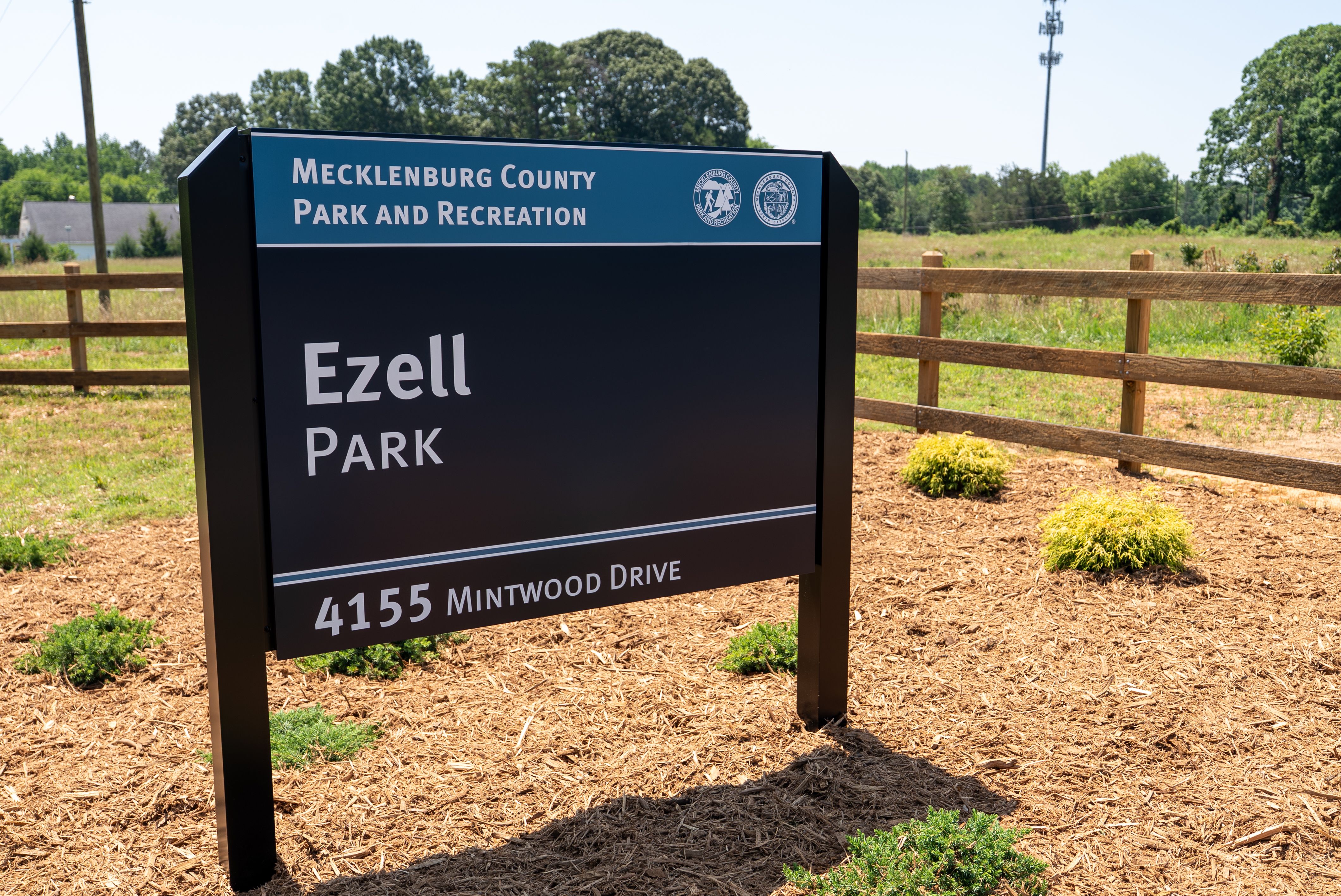 Sign reads 'Mecklenburg County Park and Recreation' on a blue panel with two logos; large 'Ezell Park' on a dark panel, showing '4155 Mintwood Drive' at bottom, with a wooden fence and grassy field behind.