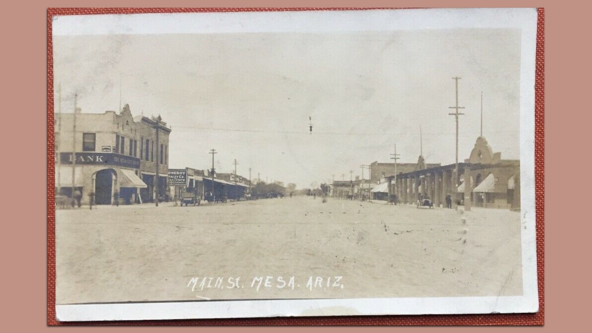 A vintage black-and-white postcard shows a wide, unpaved Main Street in downtown Mesa, Arizona, lined with two-story brick and stucco commercial buildings, storefront awnings and utility poles.
