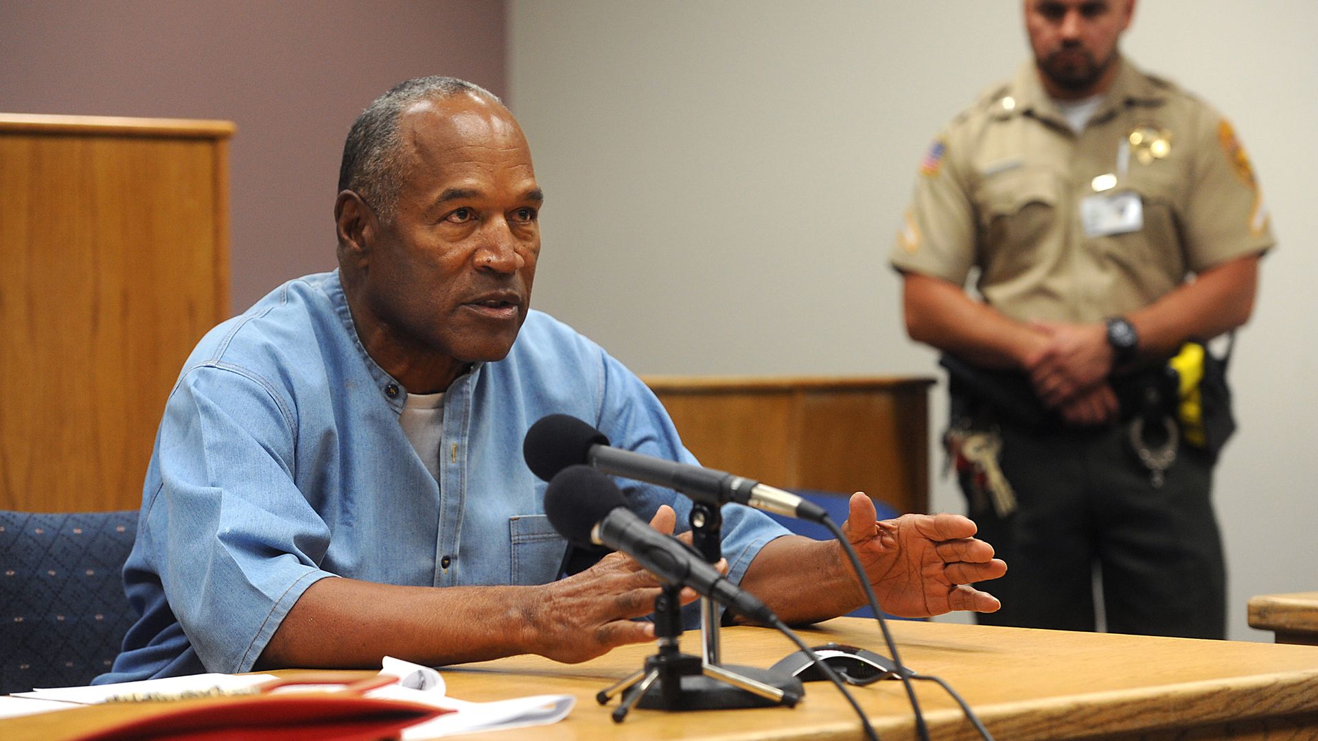 O.J. Simpson speaks seated at a table speaks into microphones. An officer stands behind him.