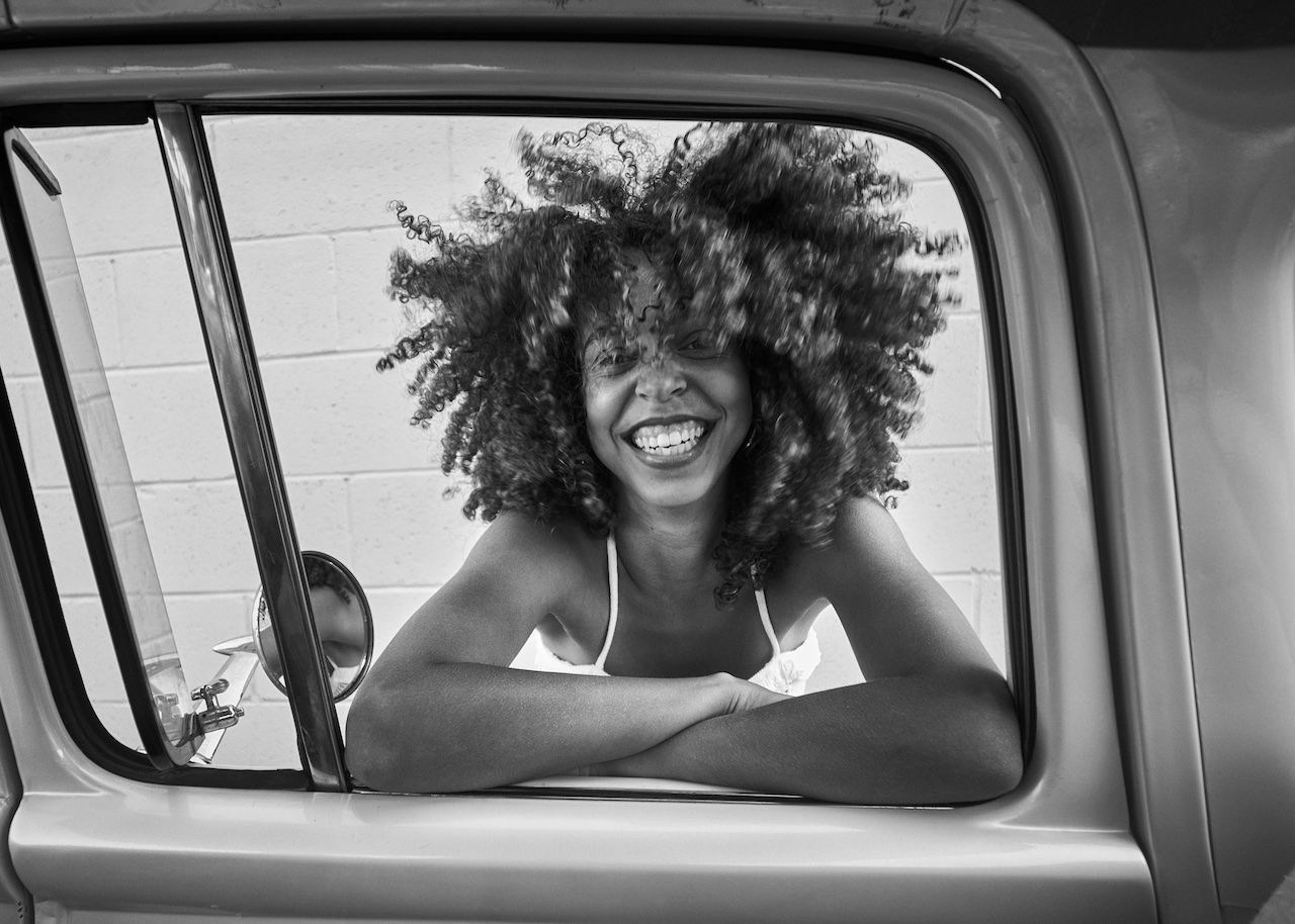 Black-and-white photo of a joyful woman with curly hair leaning through a car window, wearing a white tank top; brick wall behind her and a side mirror visible.
