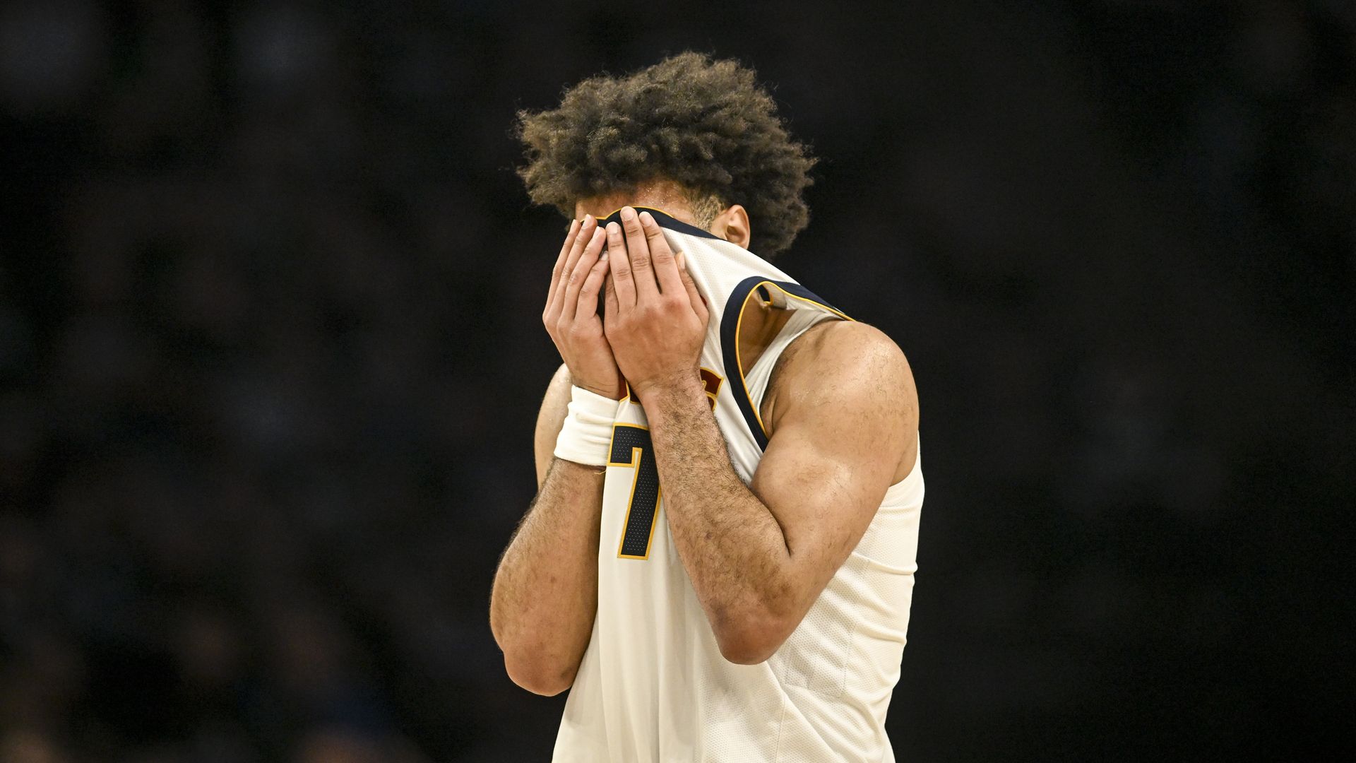 Basketball player in a white jersey covers his face with both hands, showing curly hair, against a dark, blurred arena background.