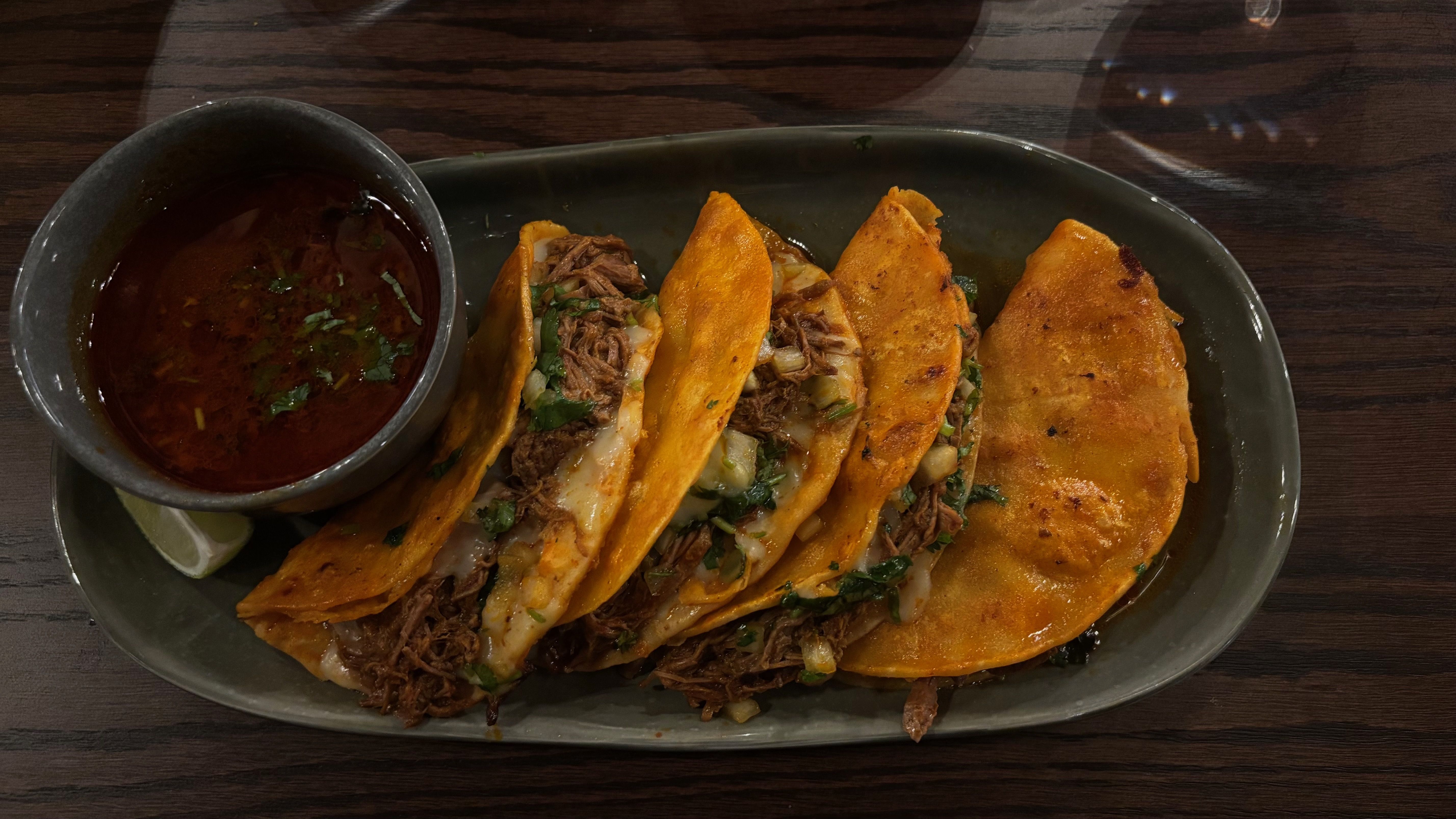 Plate of four orange-hued beef-filled tacos with chopped onions and cilantro, served with a small gray bowl of reddish dipping sauce and a wedge of lime on a dark wooden table.