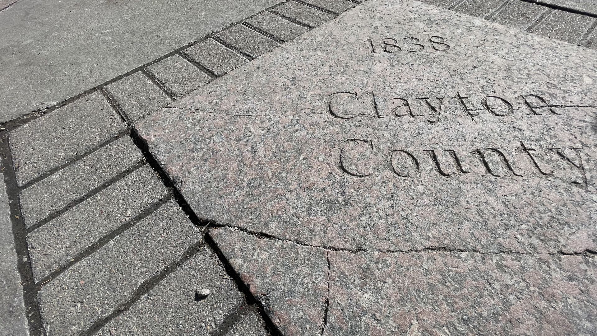 Close-up of a cracked granite plaque set in pavement, engraved with "1838 Clayton County" text in black.