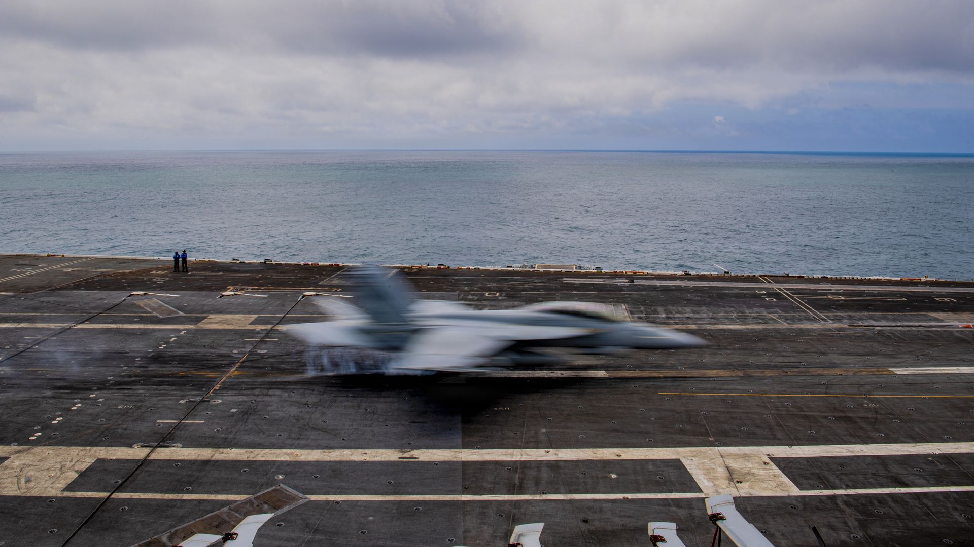 Blurry fighter jet speeds off a dark carrier deck toward the sea while two people stand at the edge watching. A cloudy sky and calm ocean stretch to the horizon.