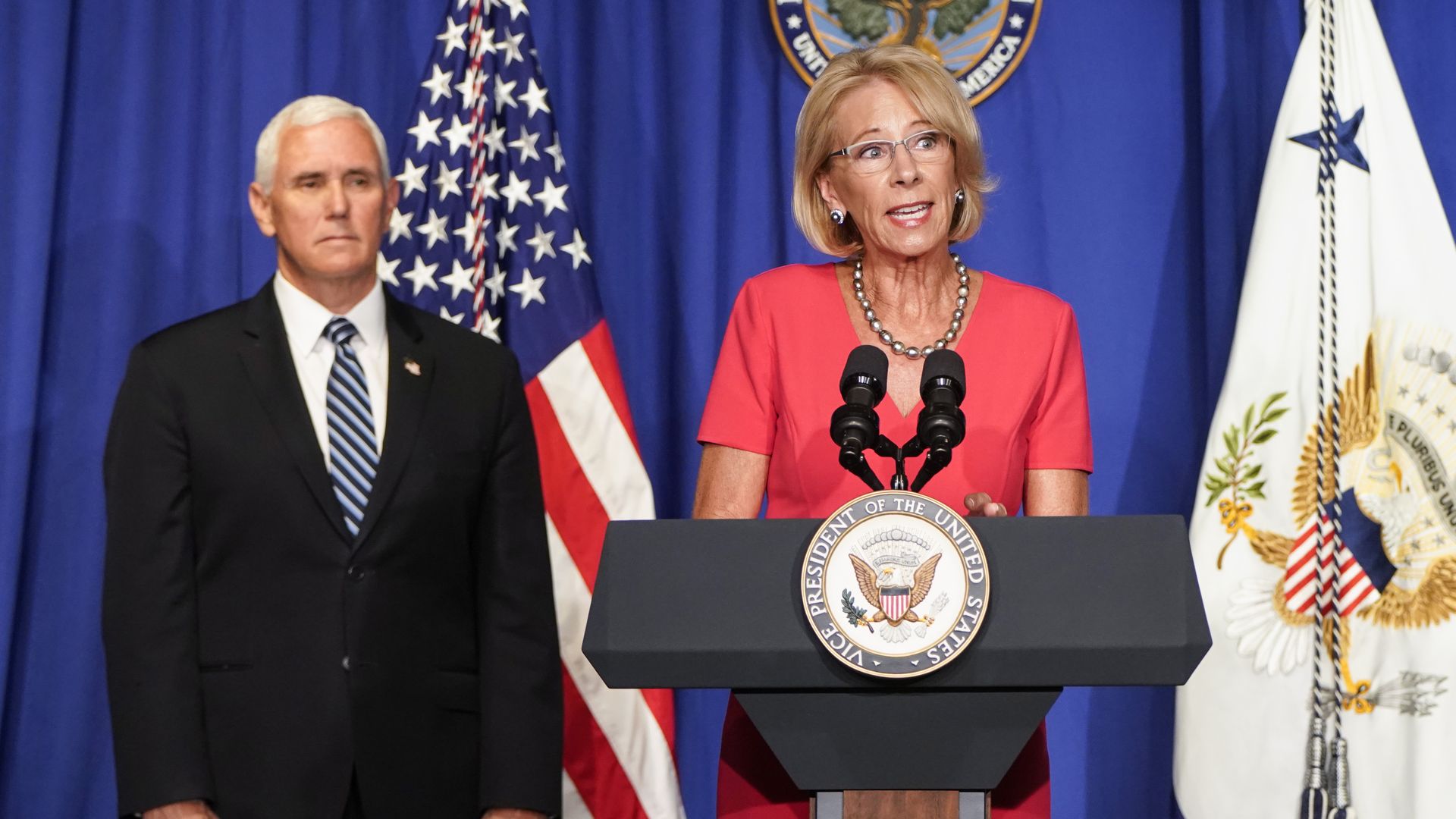 Betsy DeVos speaking at podium while Mike Pence looks on 