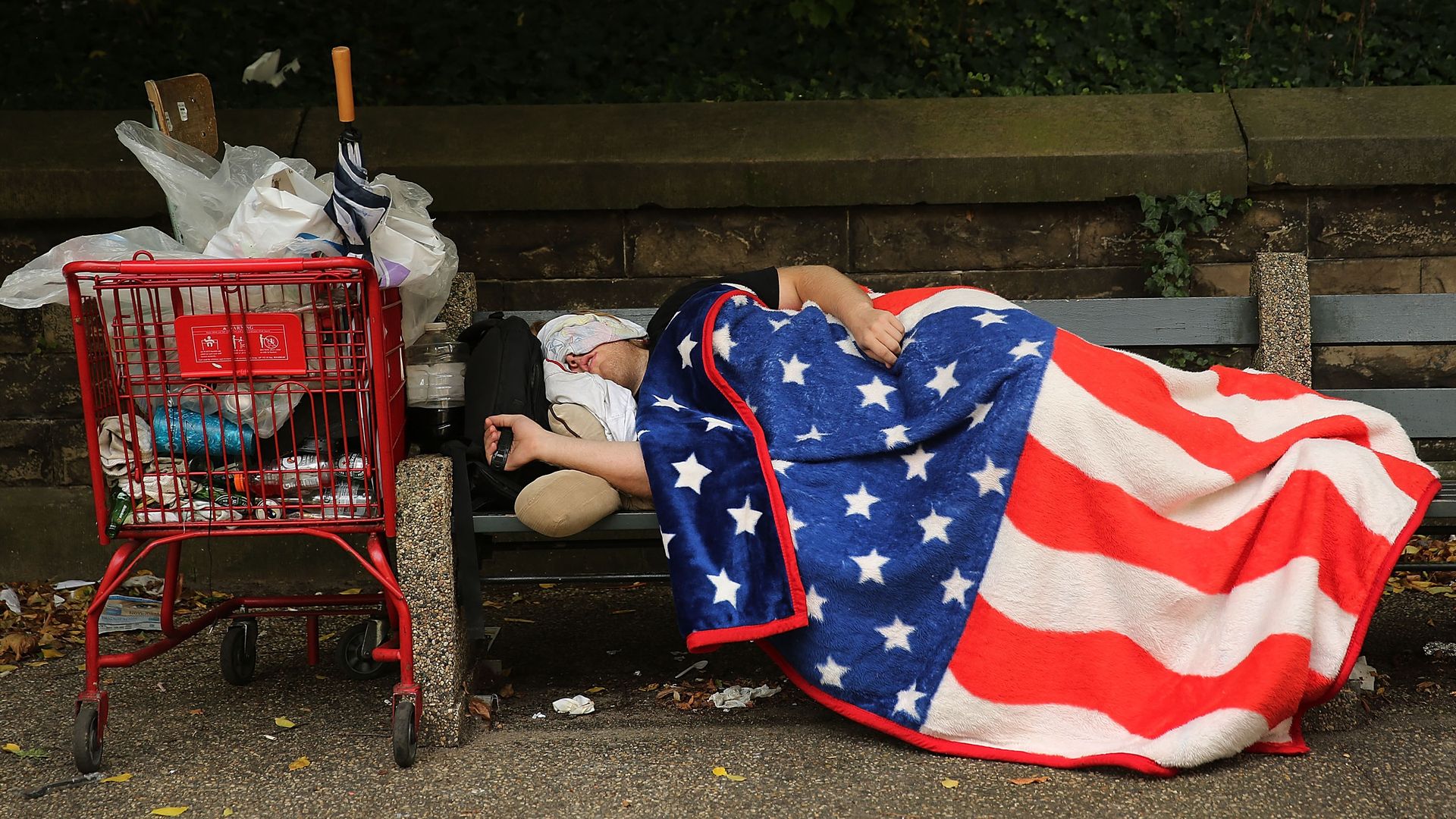 An unhoused man sleeps on a bench with an American Flag blanket draped over him, next to grocery cart