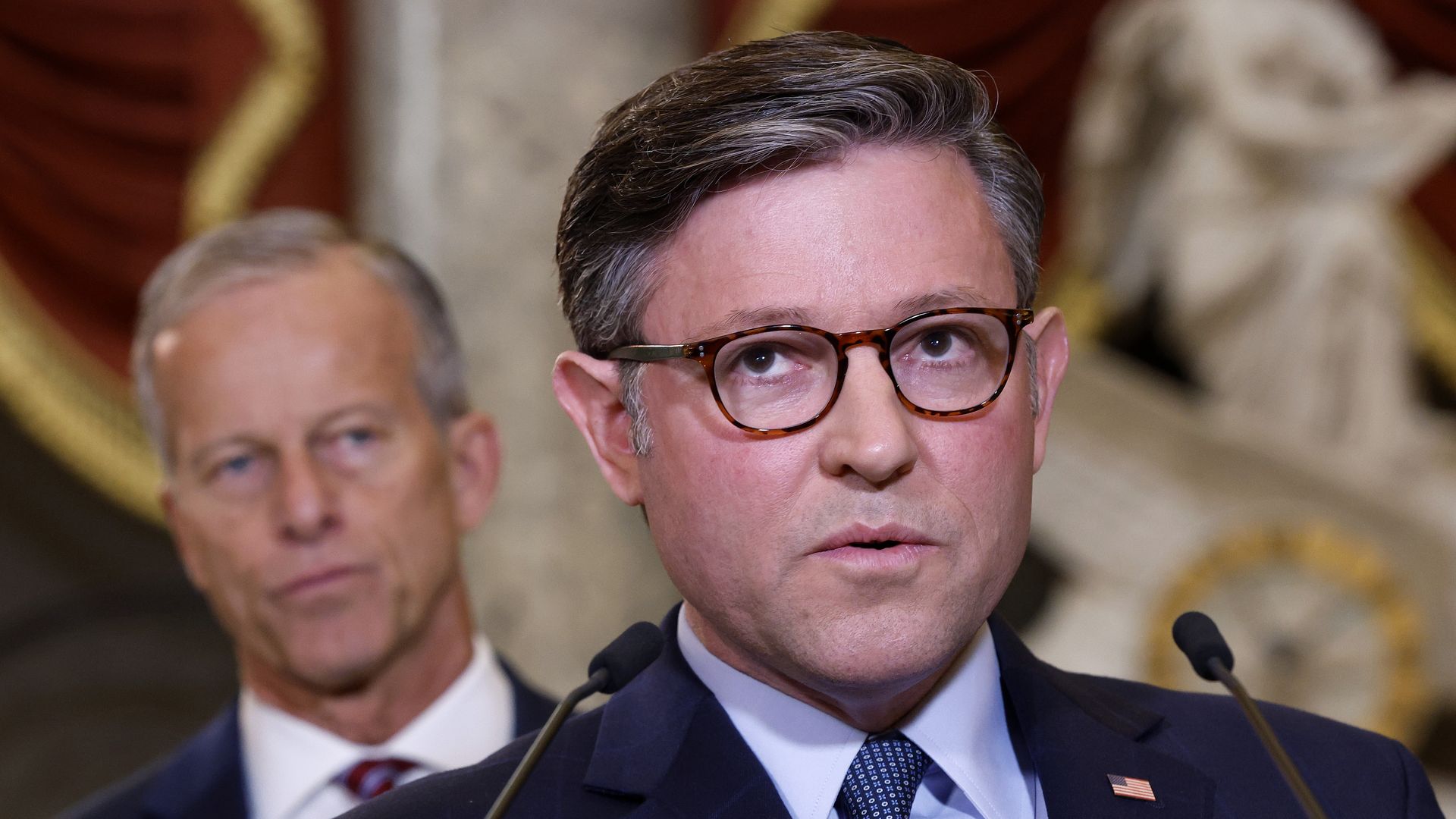 Man in dark suit and tortoiseshell glasses speaks at microphones; patterned tie; lapel pin American flag; blurred man in background, ornate backdrop.