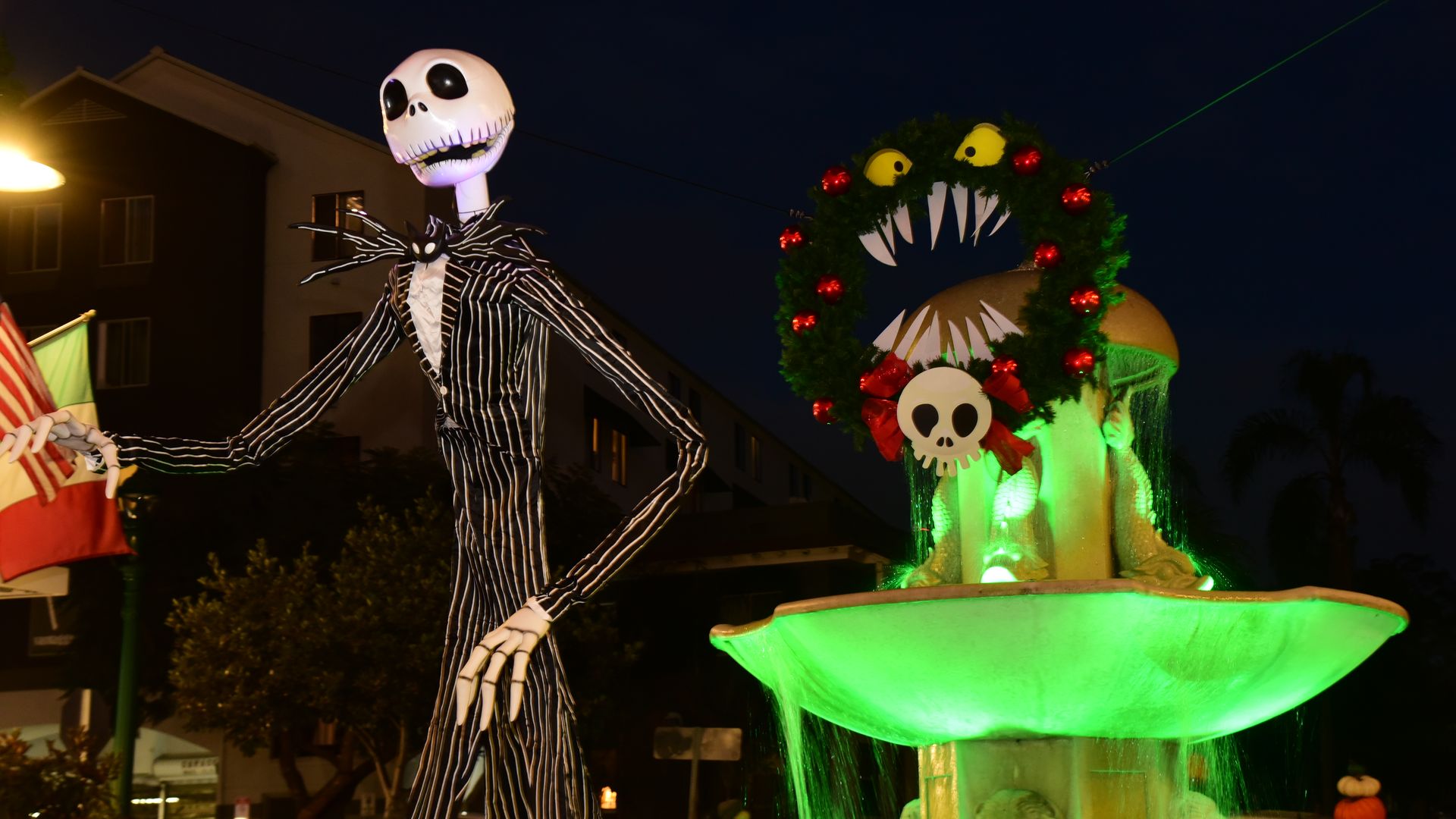 Nighttime parade float featuring a tall Jack Skellington in a pinstripe suit with long skeletal arms, beside a green glowing wreath with sharp white teeth and yellow eyes.