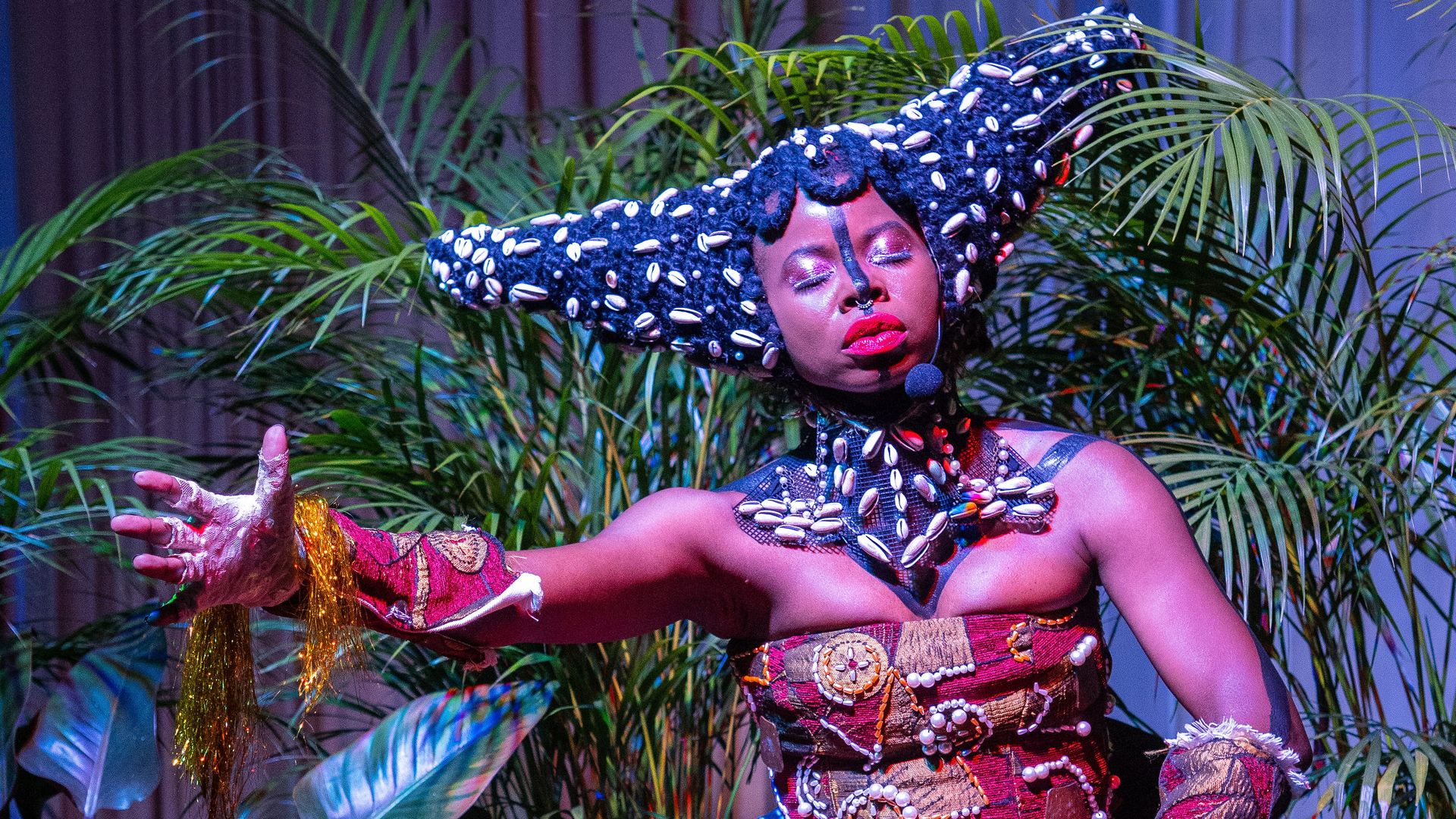 Performer in a shell-studded, horned headdress with purple makeup and red lips. Outstretched arms, red-and-gold fringe skirt, gold tassels; tropical plants and a drum in the foreground.