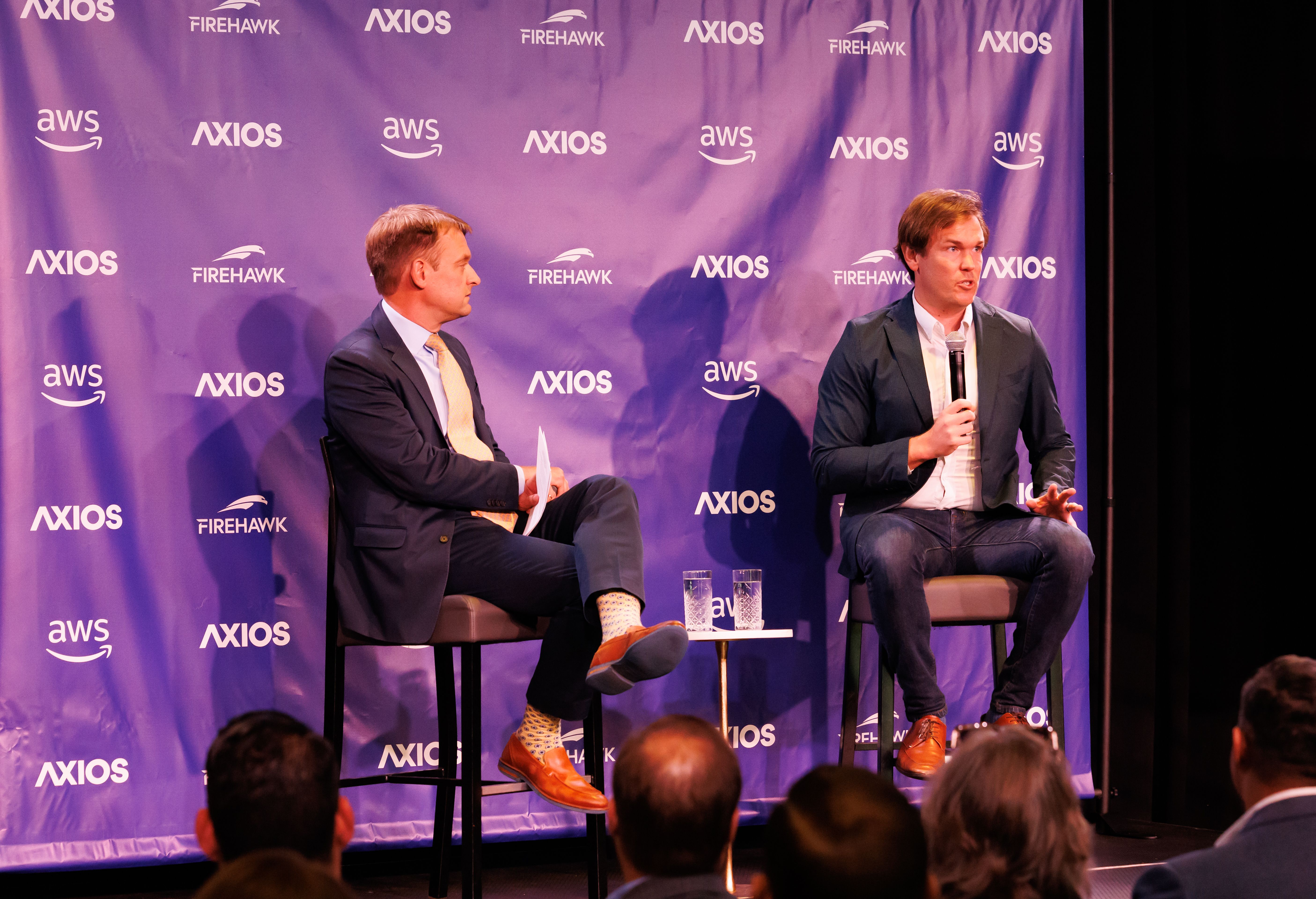 Two men in suits seated on stage at an Axios event with purple backdrop showing logos for Axios, Firehawk, and AWS. One man holds a microphone, the other holds papers.