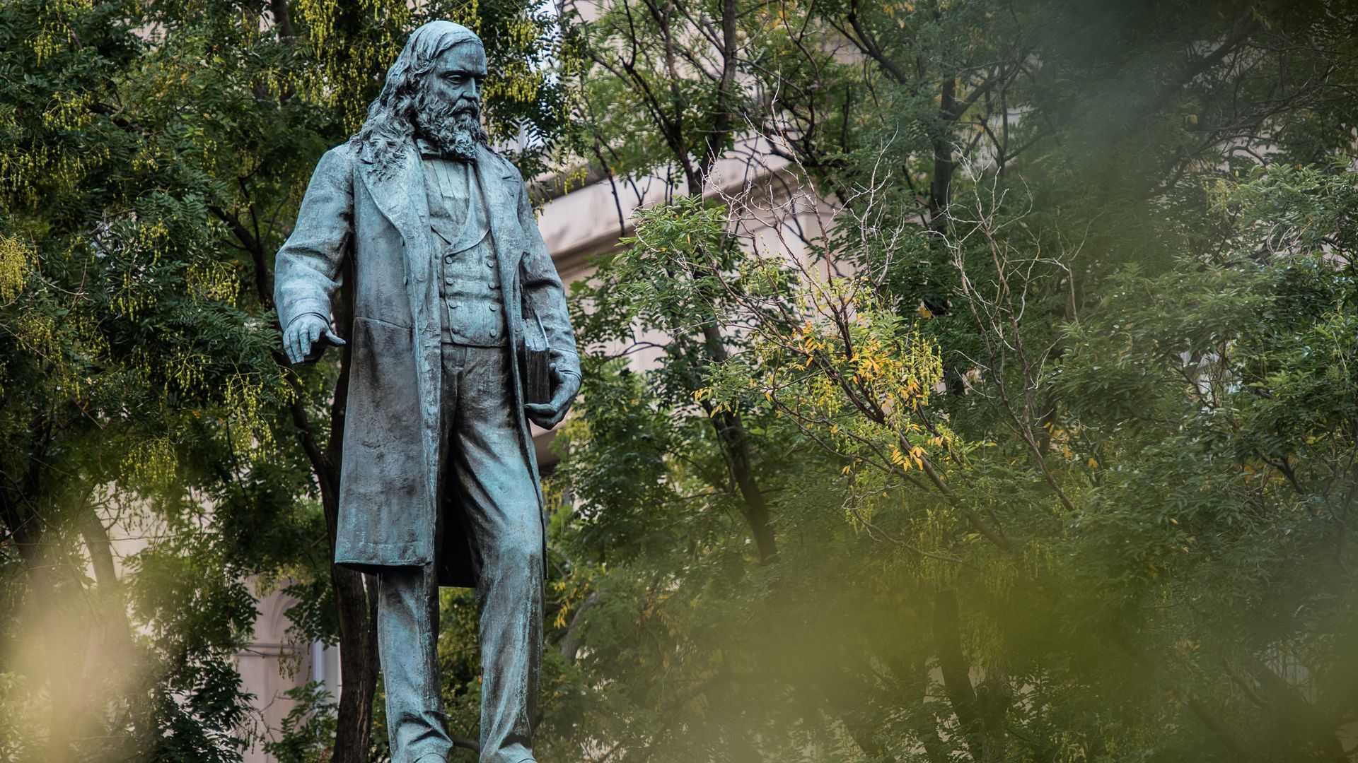 Bronze statue of a bearded man in a long coat holding a book, standing on a pedestal surrounded by green trees and a building in the background.