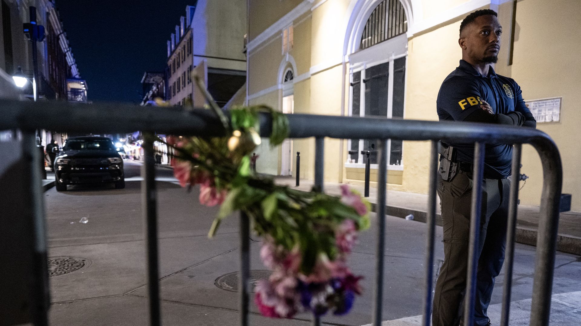 An FBI agent stands with his arms crossed behind a barricade. A bouquet of flowers is tied to the barricade.