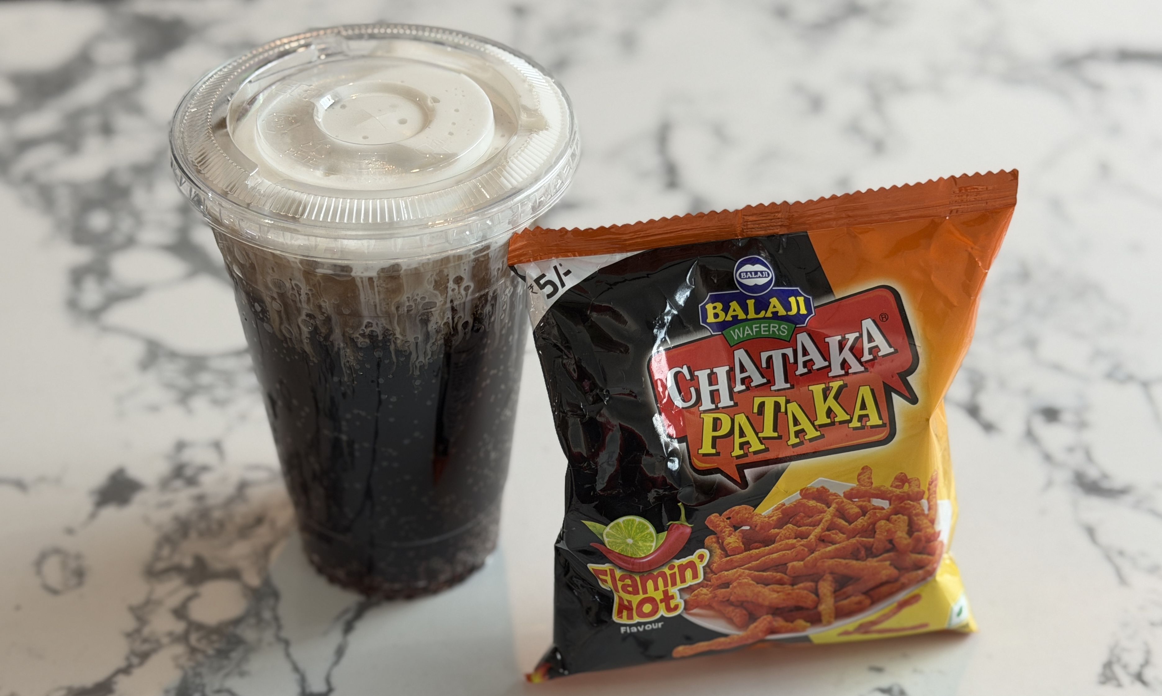 Plastic cup of dark soda with ice next to a bag of Balaji Chataka Pataka Flamin' Hot flavored snacks on a white and gray marble surface.