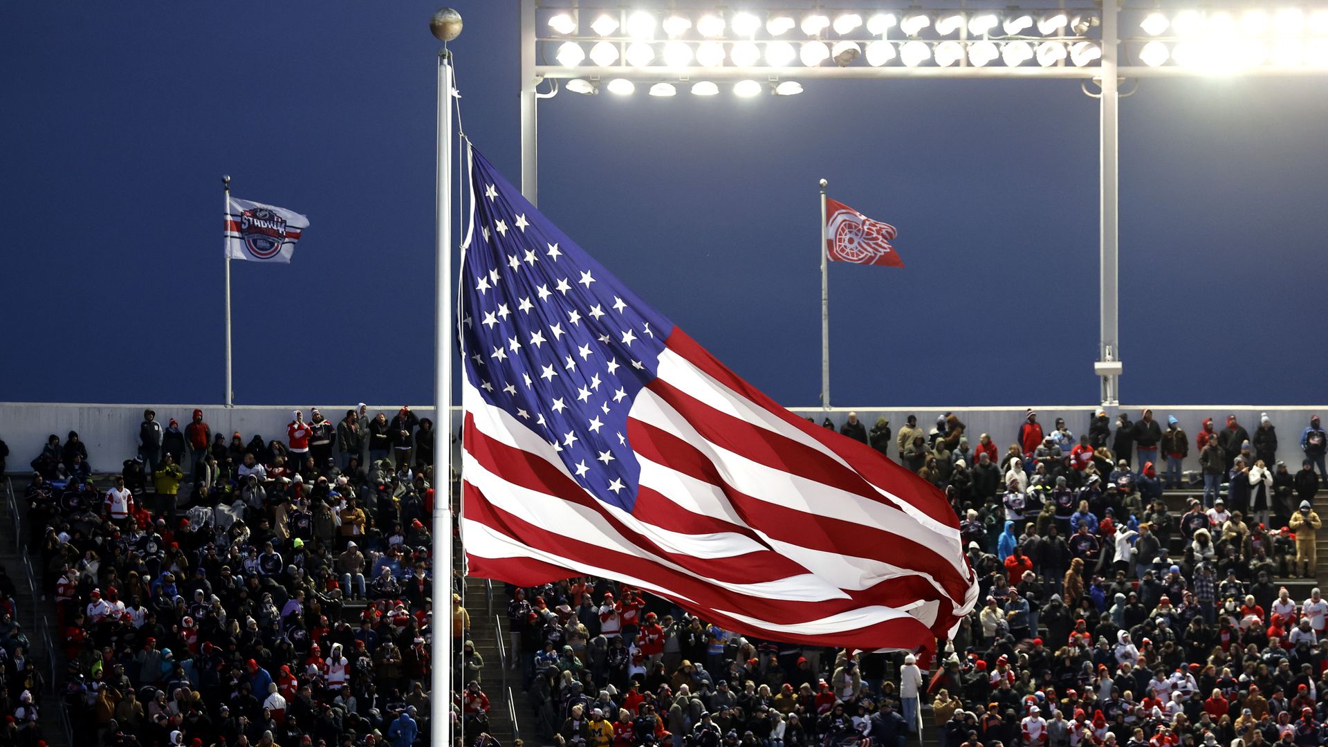 A large American flag billows over a crowded stadium, with bright floodlights overhead and smaller flags near the stands against a dark blue evening sky.