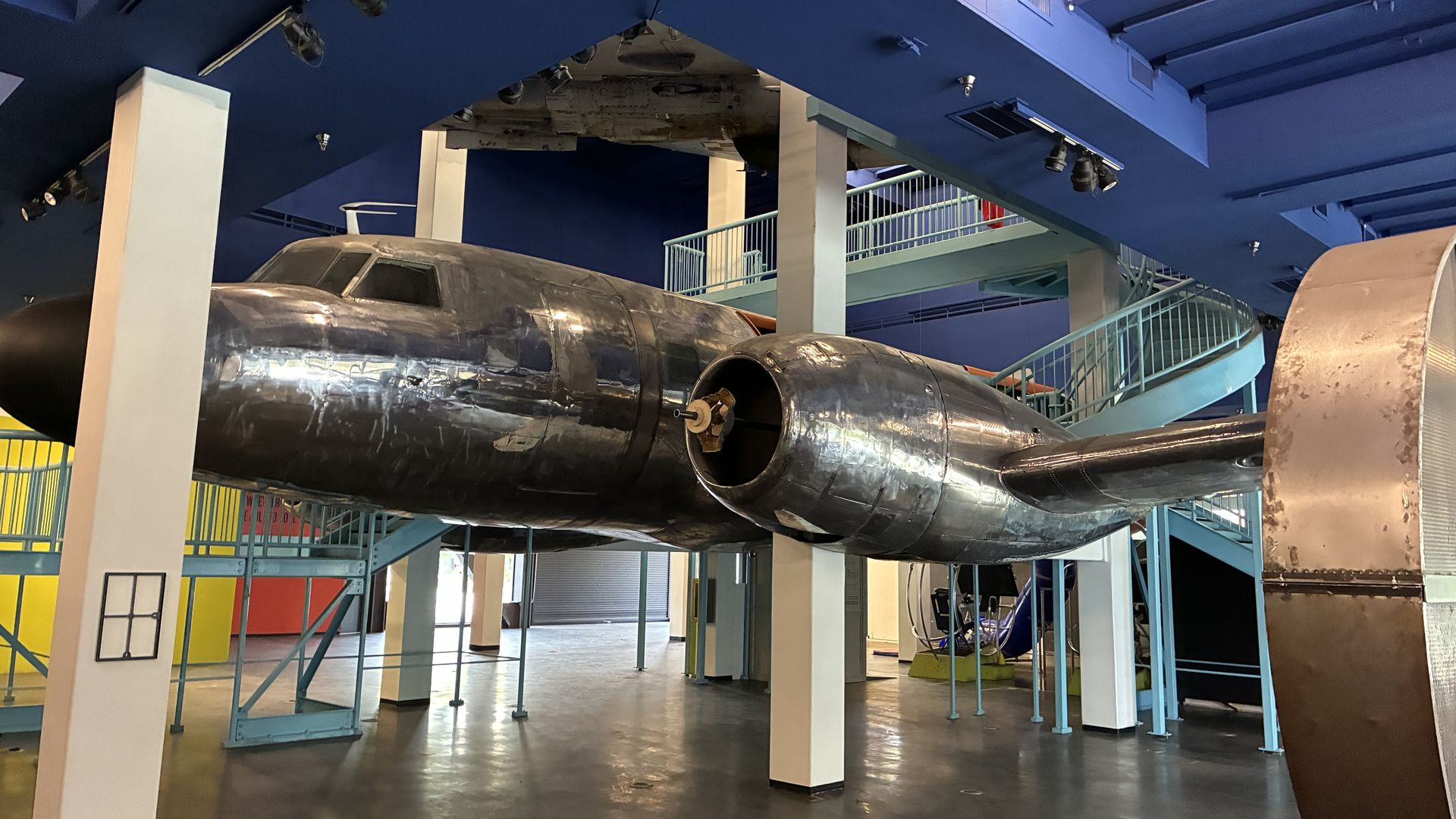 Inside a museum, a large silver vintage aircraft hangs from the ceiling. Blue structural beams, railings, and a mezzanine surround the suspended fuselage, with colorful walls in the background.