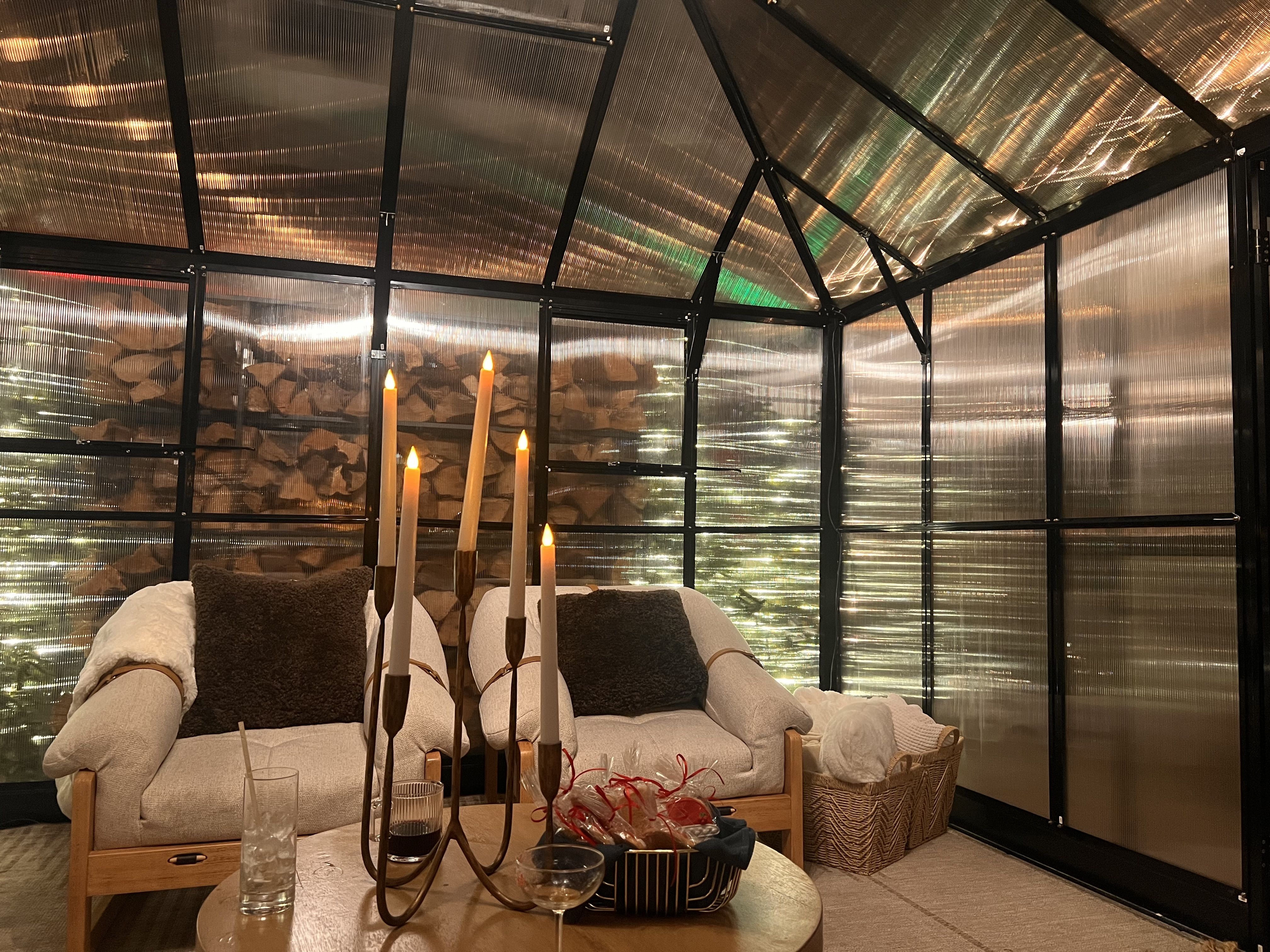 Cozy indoor seating area with two beige armchairs featuring dark cushions, a round table with lit candles and snacks, and basket with blankets inside a translucent-paneled room with wood stacked outside.