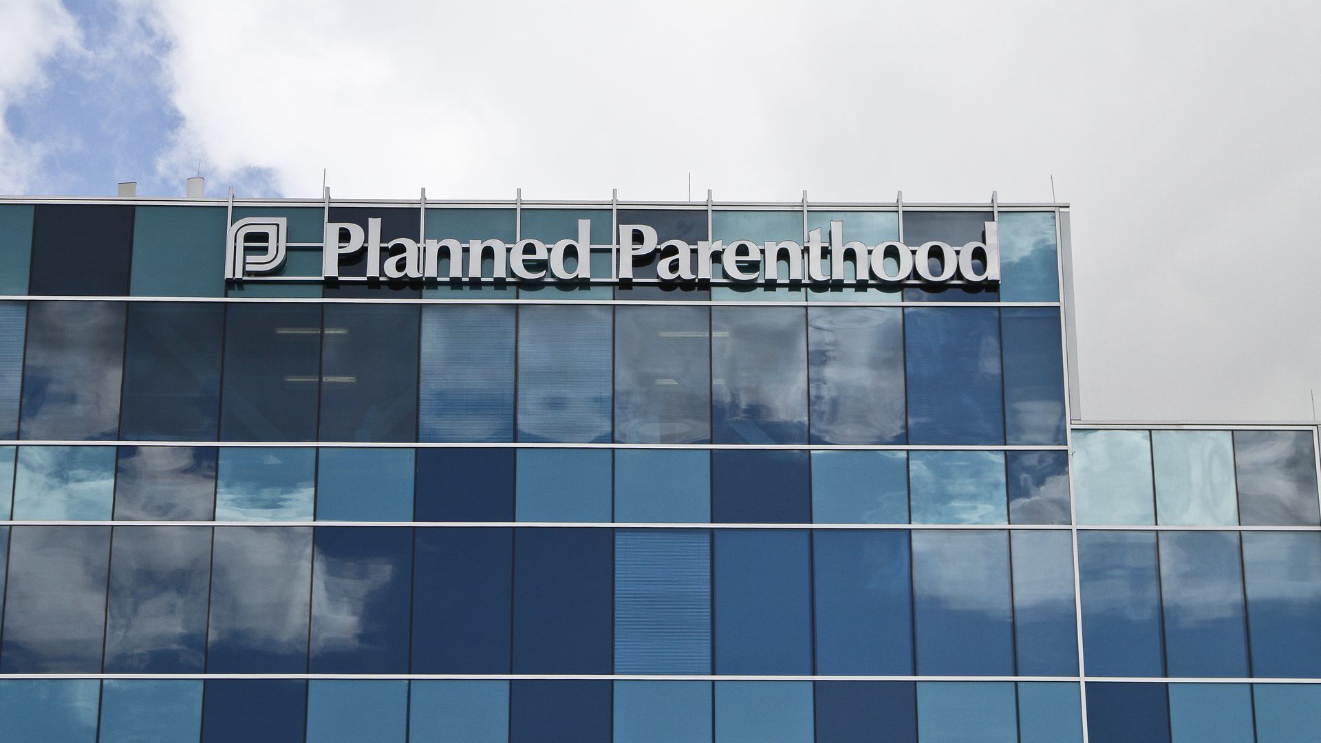 Glass building facade with blue-tinted windows reflecting the sky and clouds, featuring the white sign "Planned Parenthood" at the top.