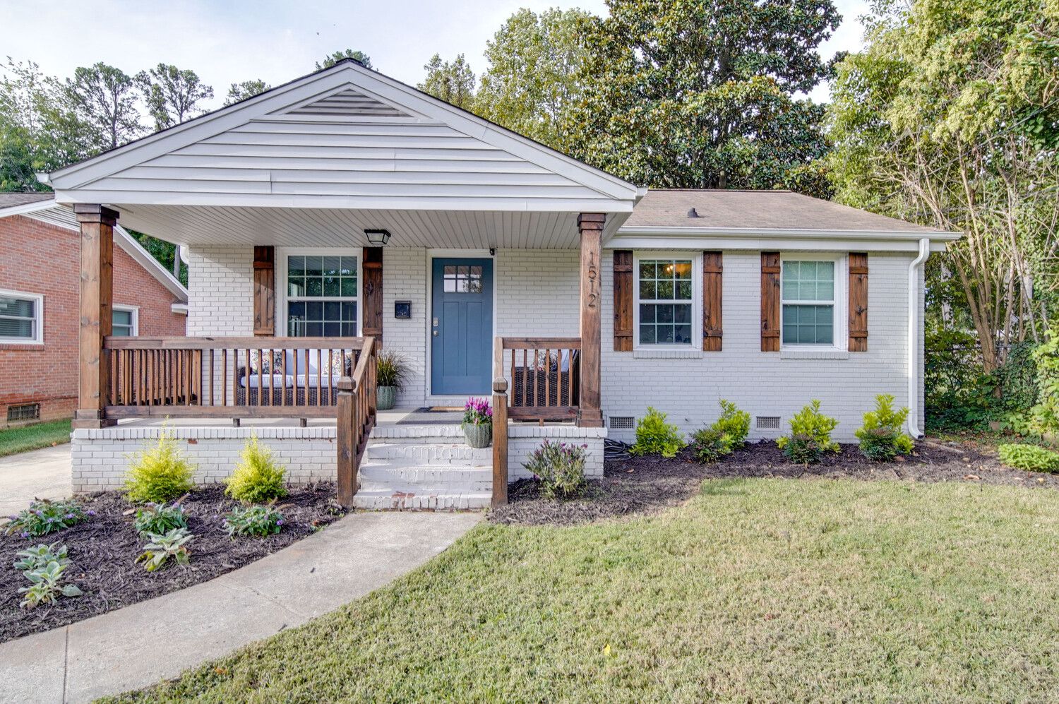 A single-story white brick house with a covered front porch, blue front door, and dark wooden columns. Windows have brown shutters; small mulched garden beds and a green lawn.