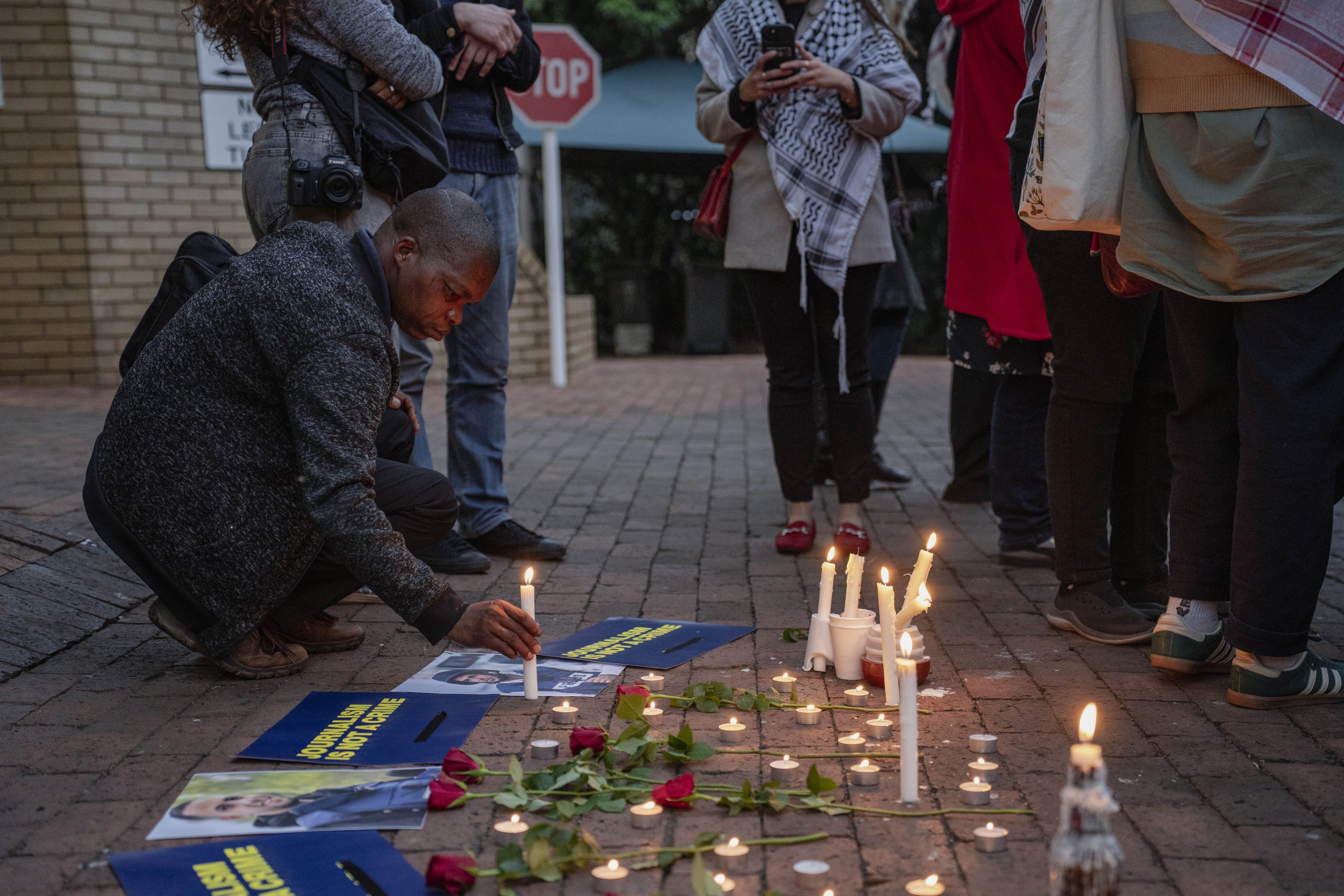 A man in a dark sweater kneels to light a candle at a vigil with roses, photos, and signs reading "JOURNALISM IS NOT A CRIME" on a brick pavement surrounded by standing people.