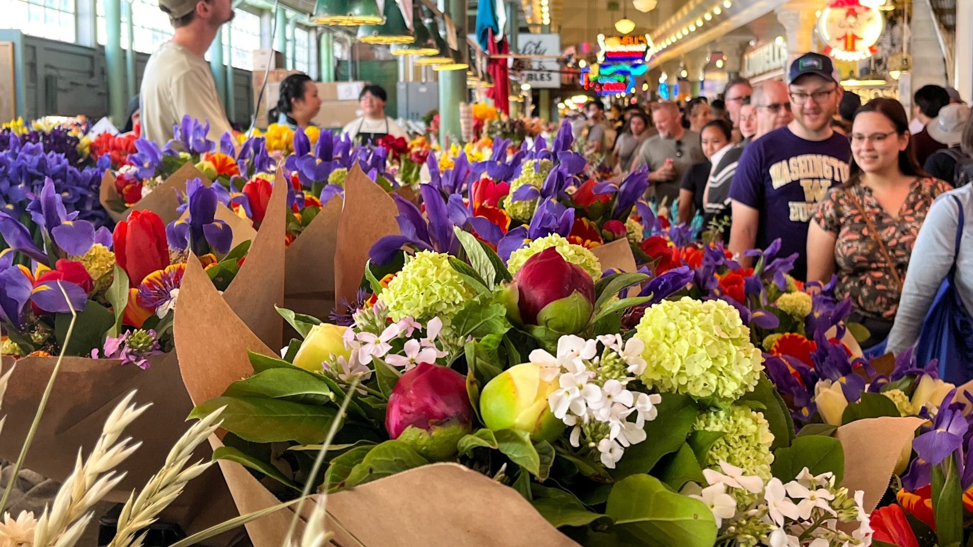 Brightly colored bouquets line stalls at Pike Place Market while people look at them as they walk by through the interior market building.