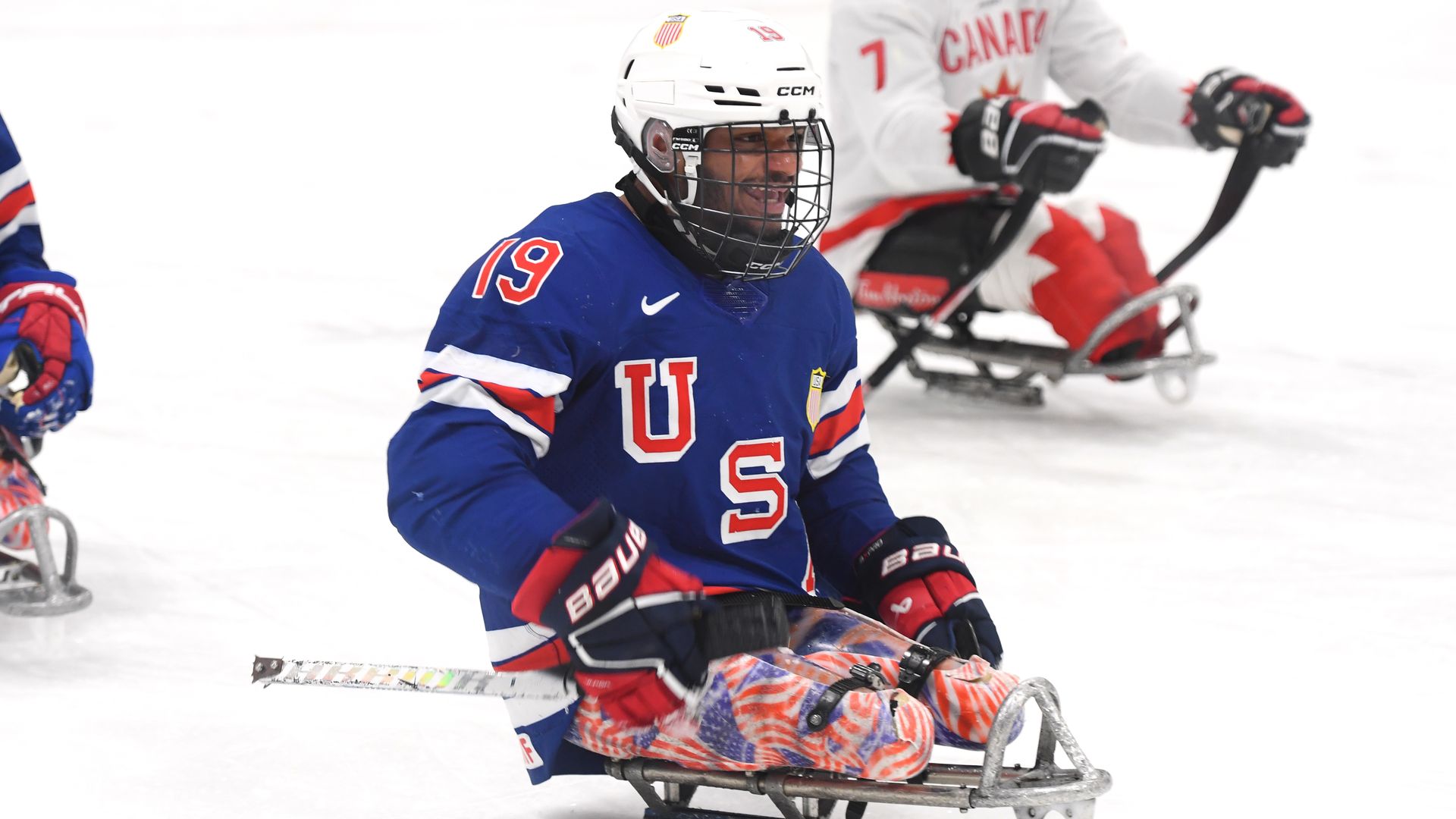 Two sledge hockey players on ice; one in a blue USA jersey, white helmet, and red gloves, the other in a white Canada jersey with red gloves and gear.