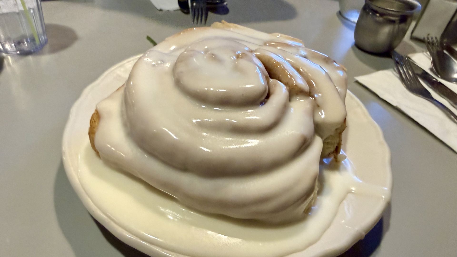 A cinnamon roll glazed with thick white icing sits on a white plate; a blurred fork, napkin, and mug appear in the background.