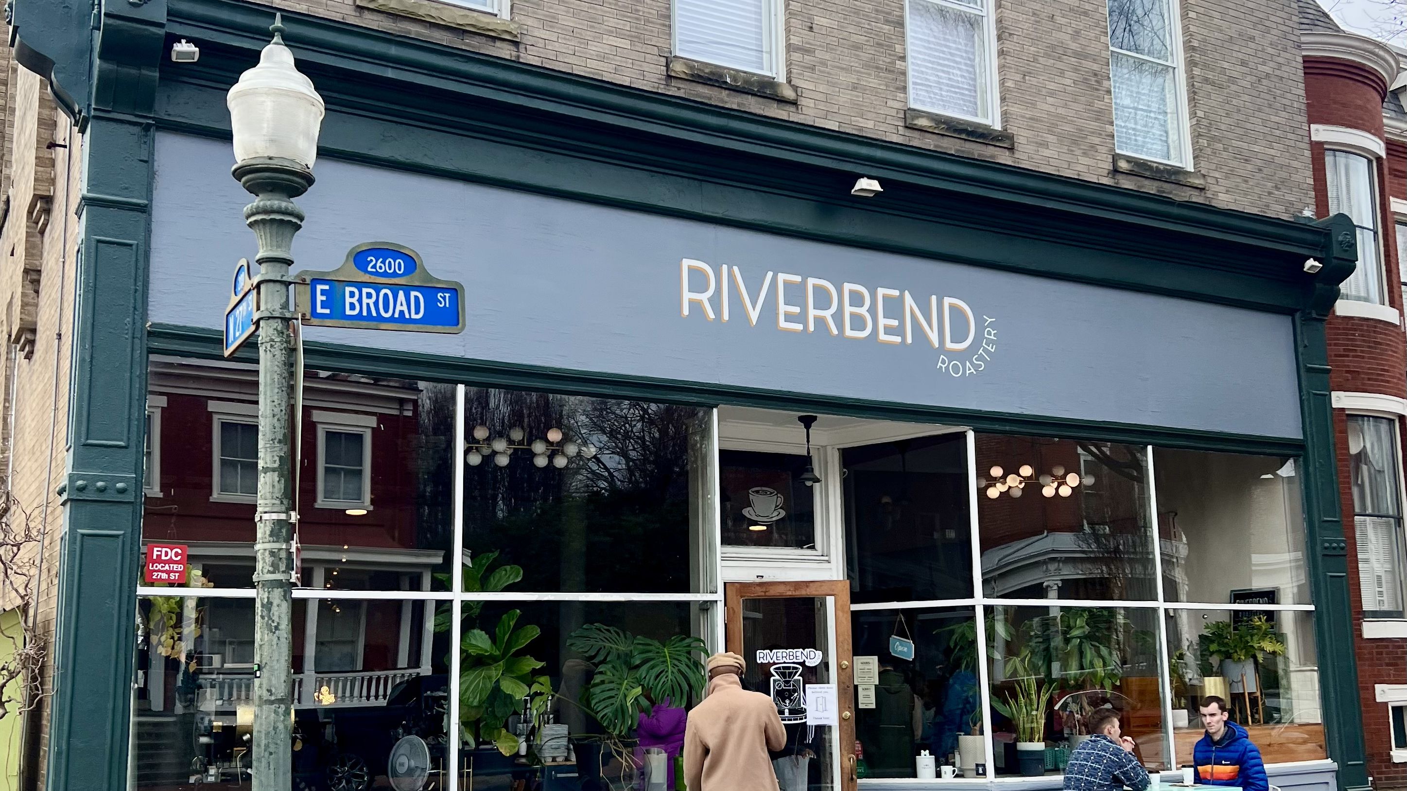A pic of a coffee shop with the words "RIVERBEND" in white letters and people sitting down outside. The windows are covered with plants.