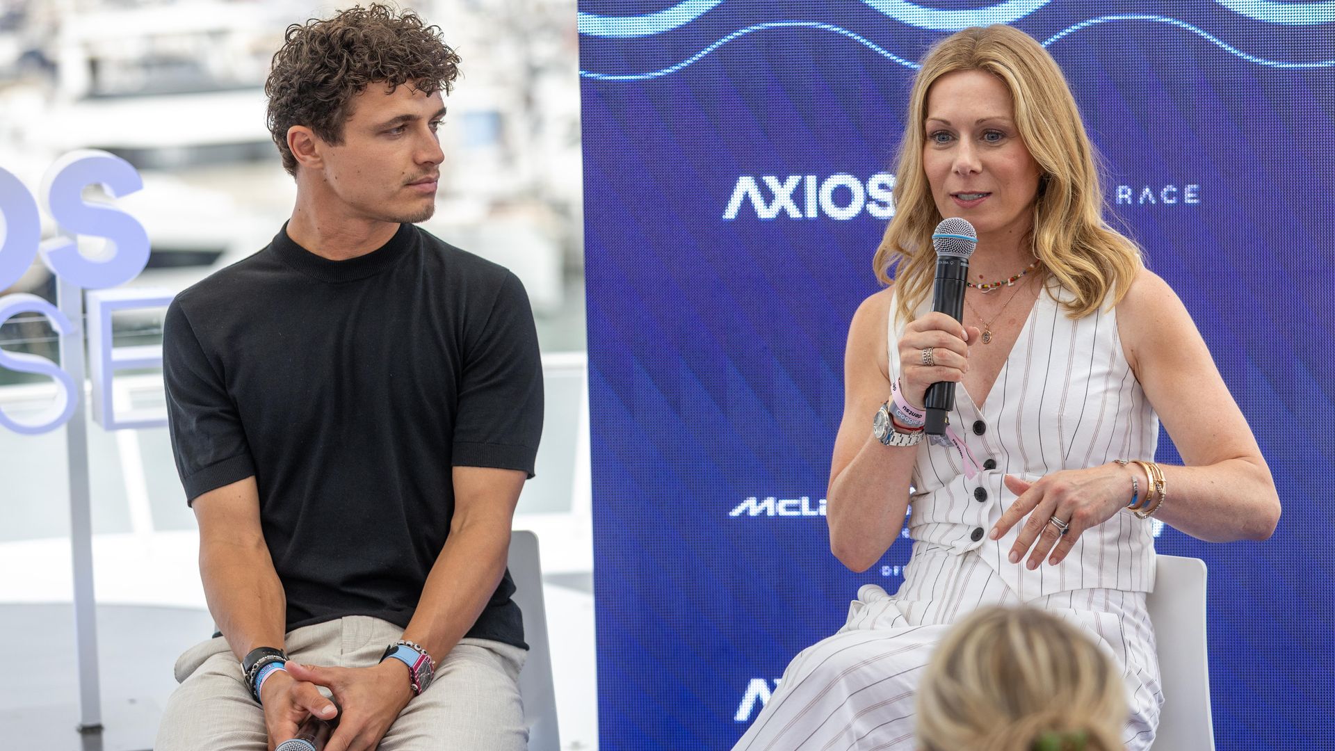 Panel scene with a man on the left in a black shirt looking toward a speaking blonde woman on the right, who holds a microphone, with a blue wavy backdrop behind them.