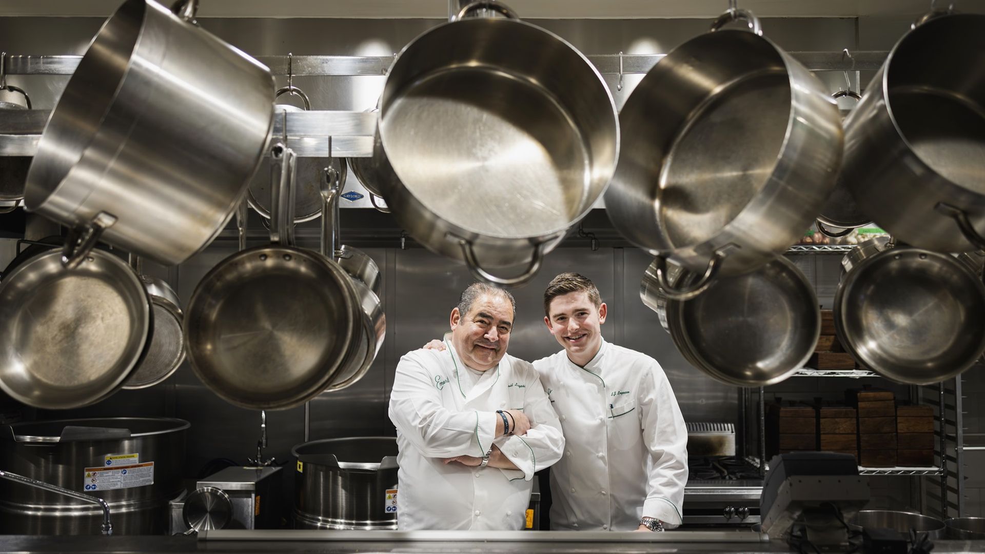 Emeril Lagasse poses for a photo with his son, E.J. The pair lean toward each other in the center of the frame, and they stand in a kitchen where pots hang from the ceiling, framing the pair.