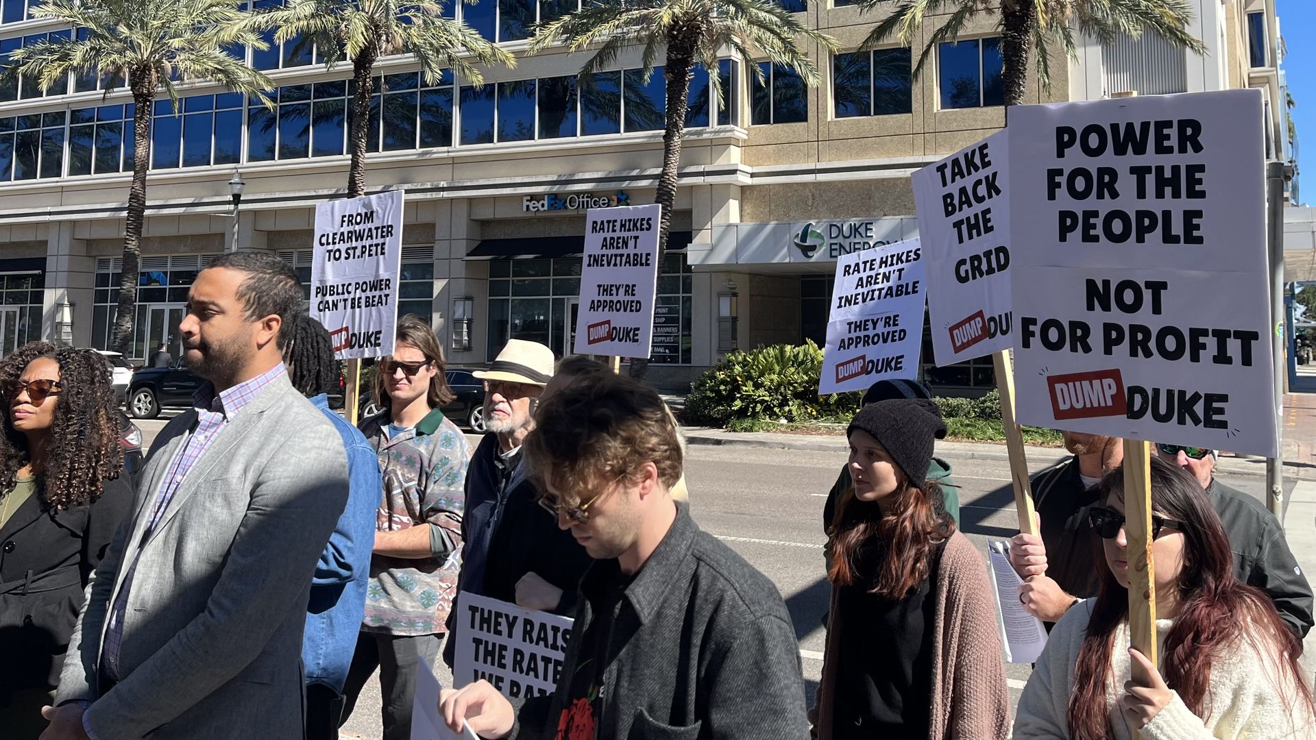 Group of protesters outside a Duke Energy office holding signs opposing Duke Energy rate hikes and advocating for public power, with palm trees and a sunny city backdrop.