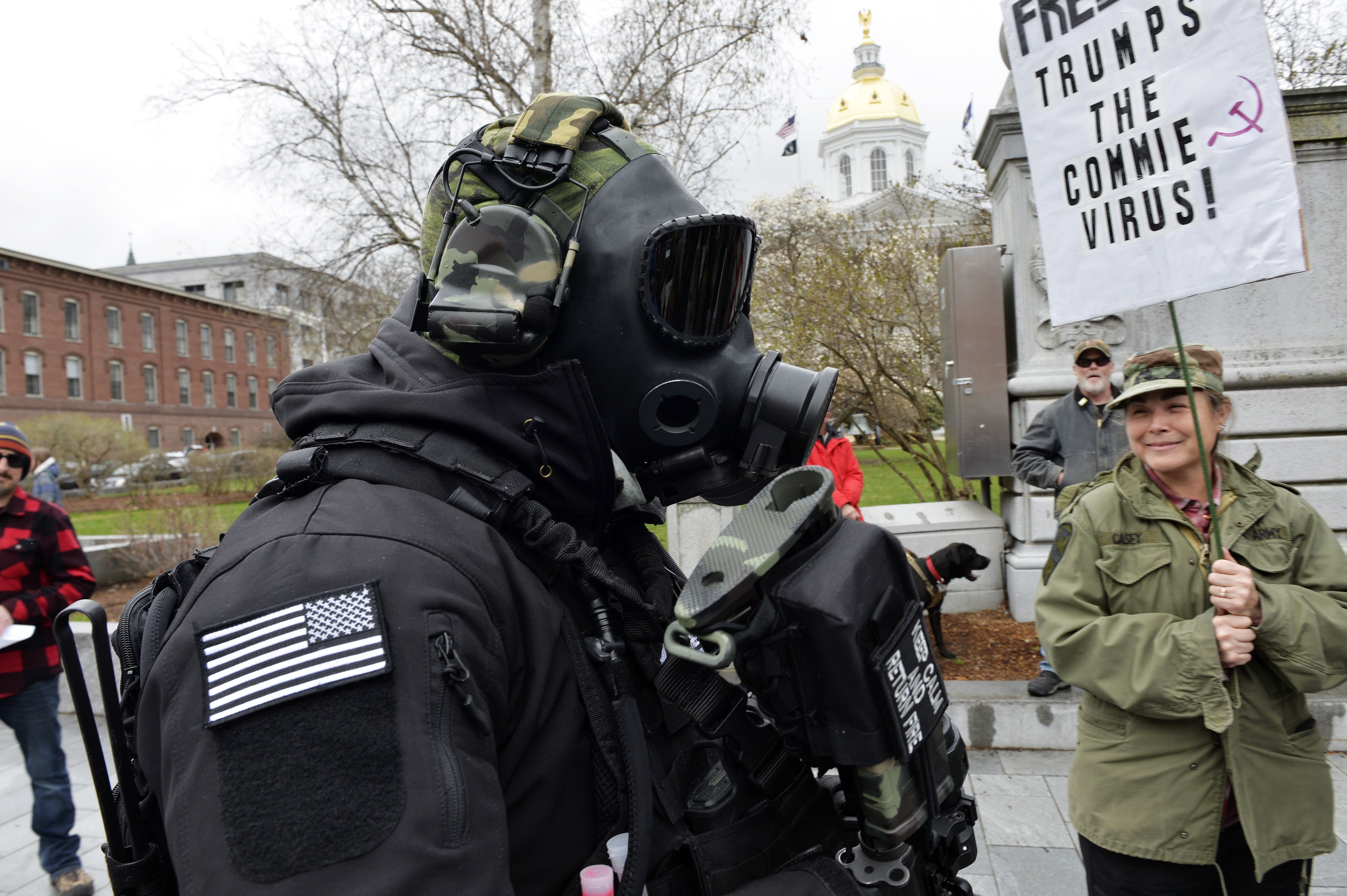 In this image, a masked and armed man walks past a sign that reads "Fear trumps the commie virus"