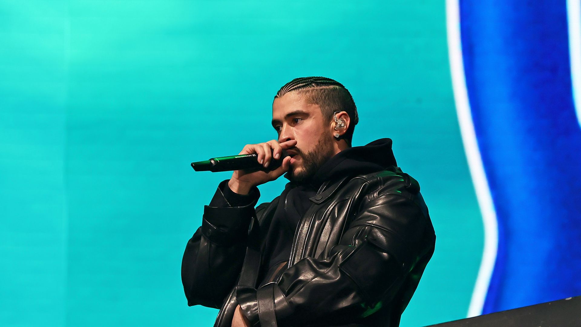 Man with braided hair and beard wearing black leather jacket and hoodie holds a microphone close to his mouth, performing on stage with teal and blue background.