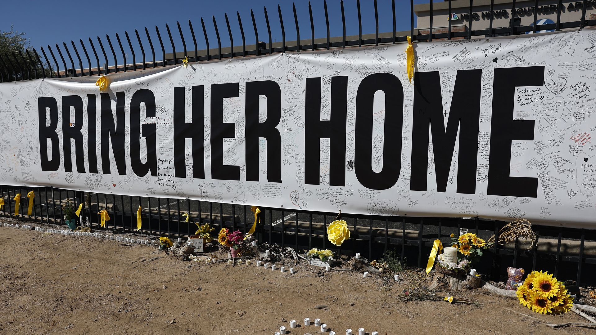 White banner with bold black text "BRING HER HOME" on a black fence. Yellow ribbons adorn the fence; memorial flowers, sunflowers, and a heart-shaped circle of candles rest on the dirt beneath.