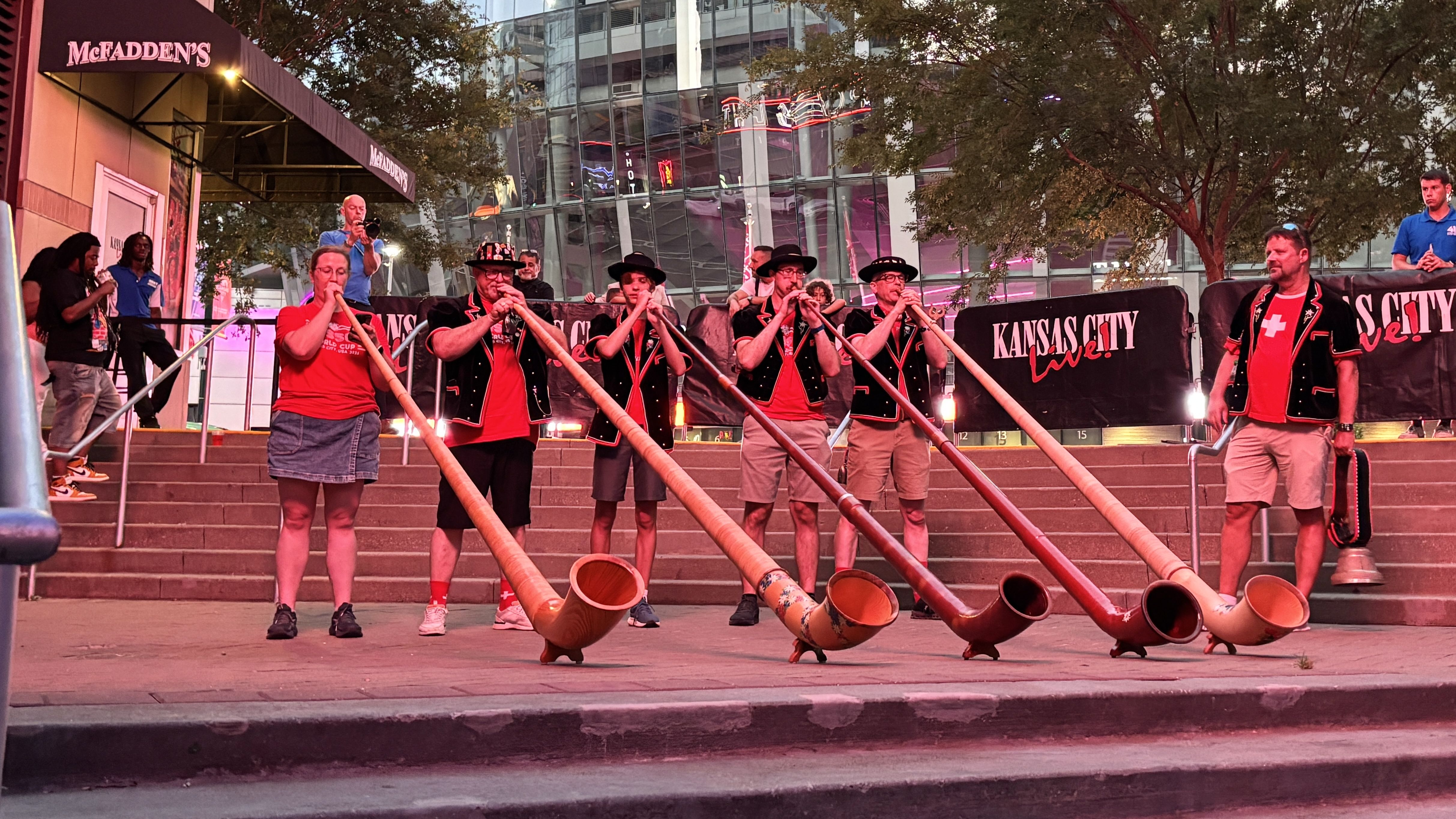 Men in red shirts and black vests play long wooden horns on steps; a Kansas City Live banner behind them, with a glass-front building and McFadden's sign to the left.