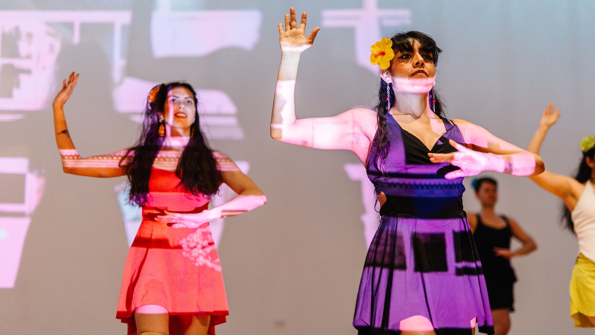 Two female dancers perform on a stage in bright dresses, one in purple with a yellow flower in her hair and the other in pink. Colorful light projections sweep the backdrop.
