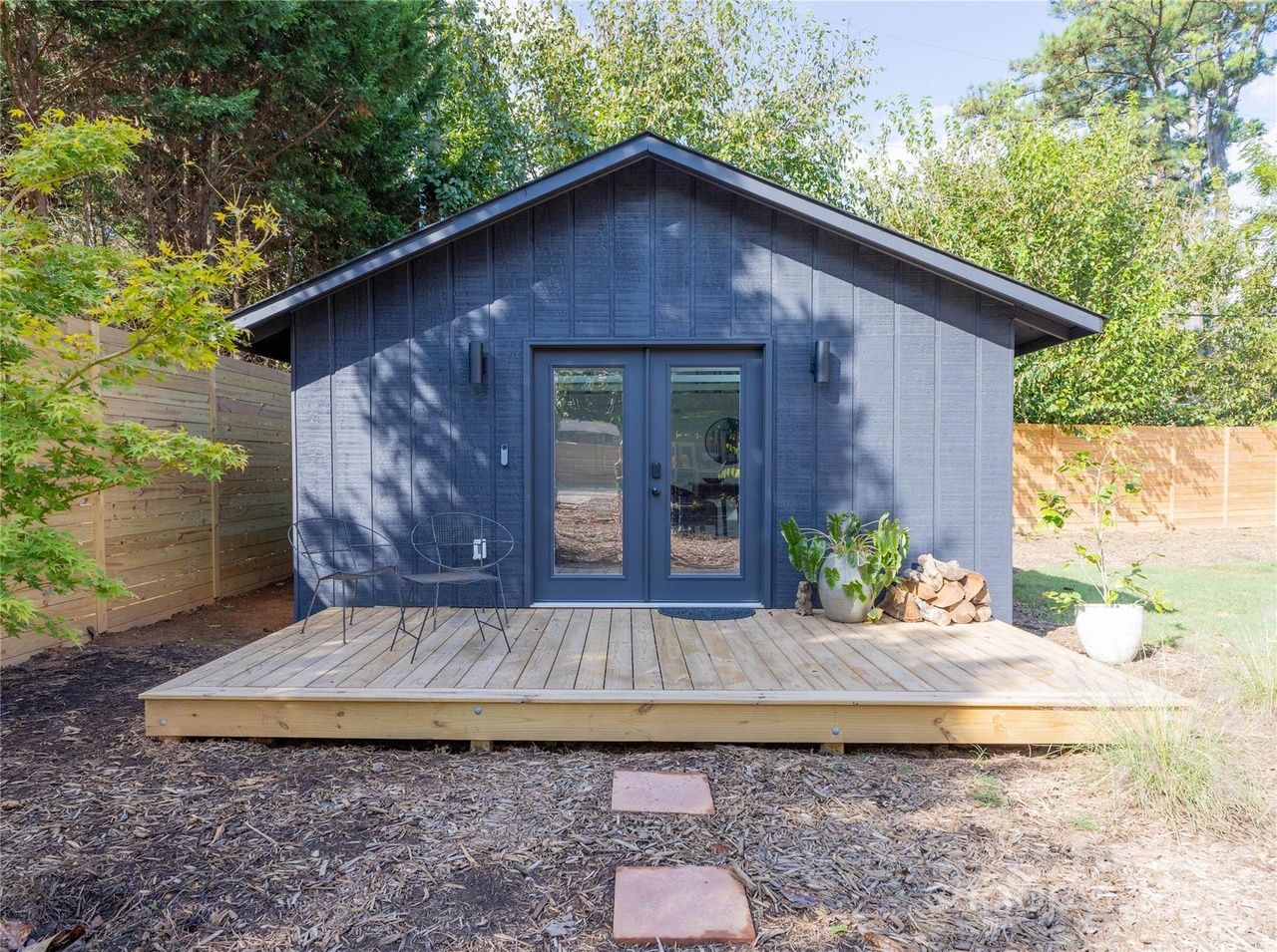 Small dark gray wooden cabin with double glass doors, two black wire chairs, potted plants, stacked firewood, and a light wooden deck surrounded by trees and a wooden fence.