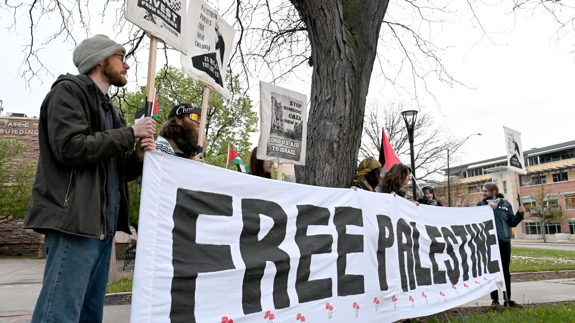 Group of protesters holding a large white banner reading "FREE PALESTINE" with red flowers, standing near a tree and holding signs with messages like "Divest from Genocide" and "Stop Bombing Gaza Now".