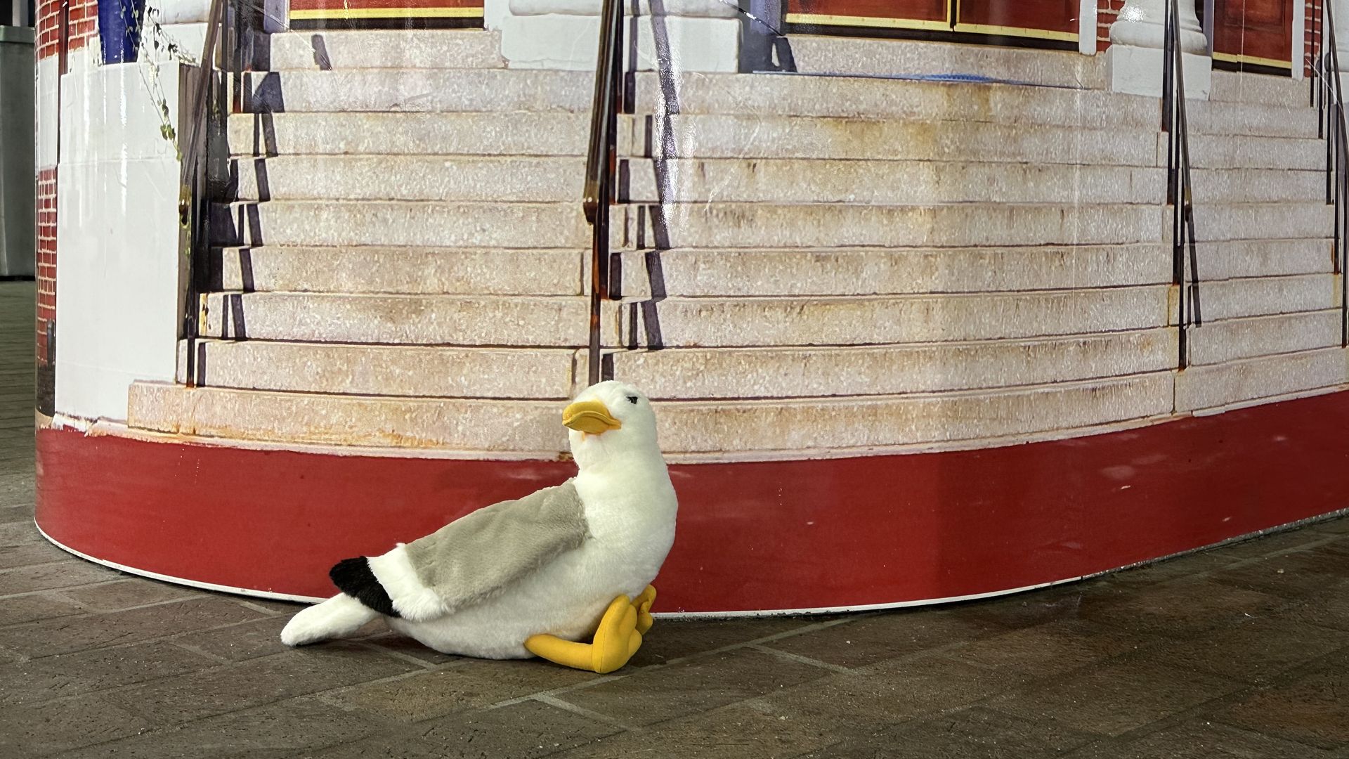 Townie, Axios Boston's plush gullscot, is perched on a concrete floor in front of a booth with an image of steps. 