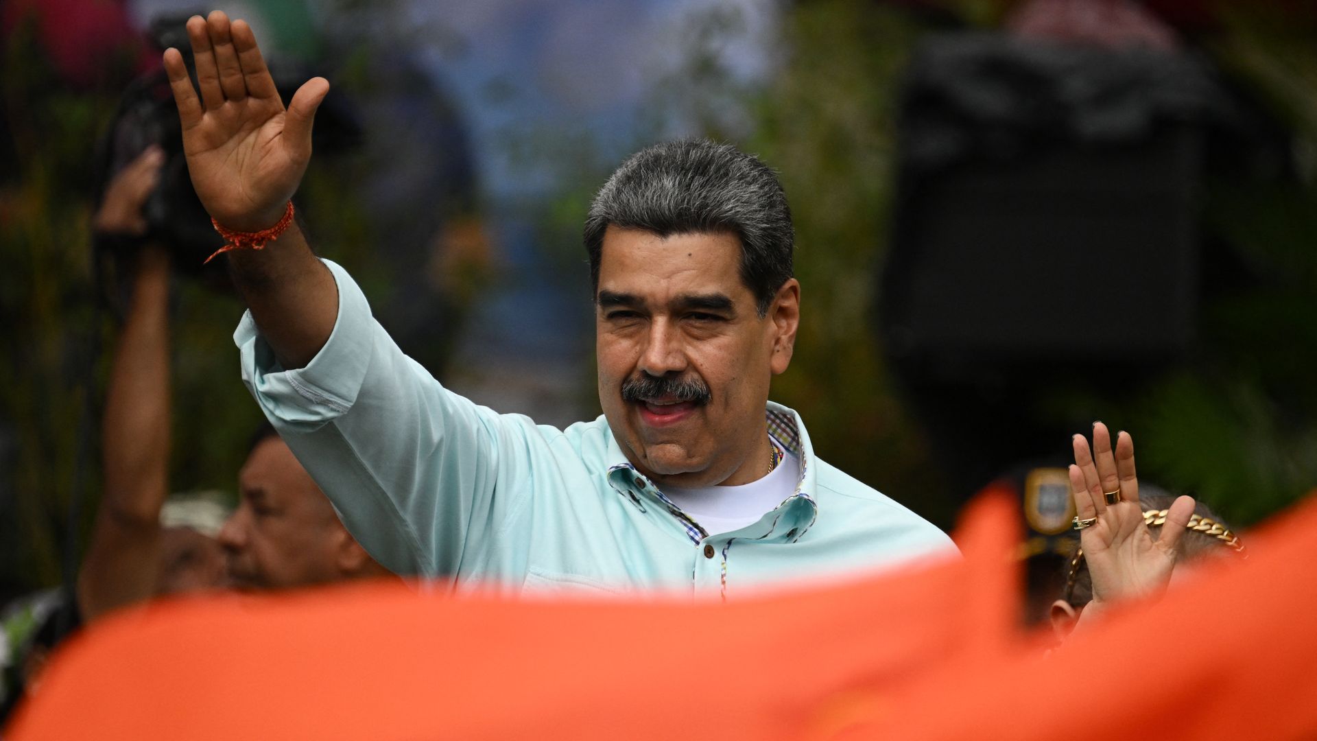 Nicolas Maduro, in an open-collared shirt, waves to a crowd with his right hand. 